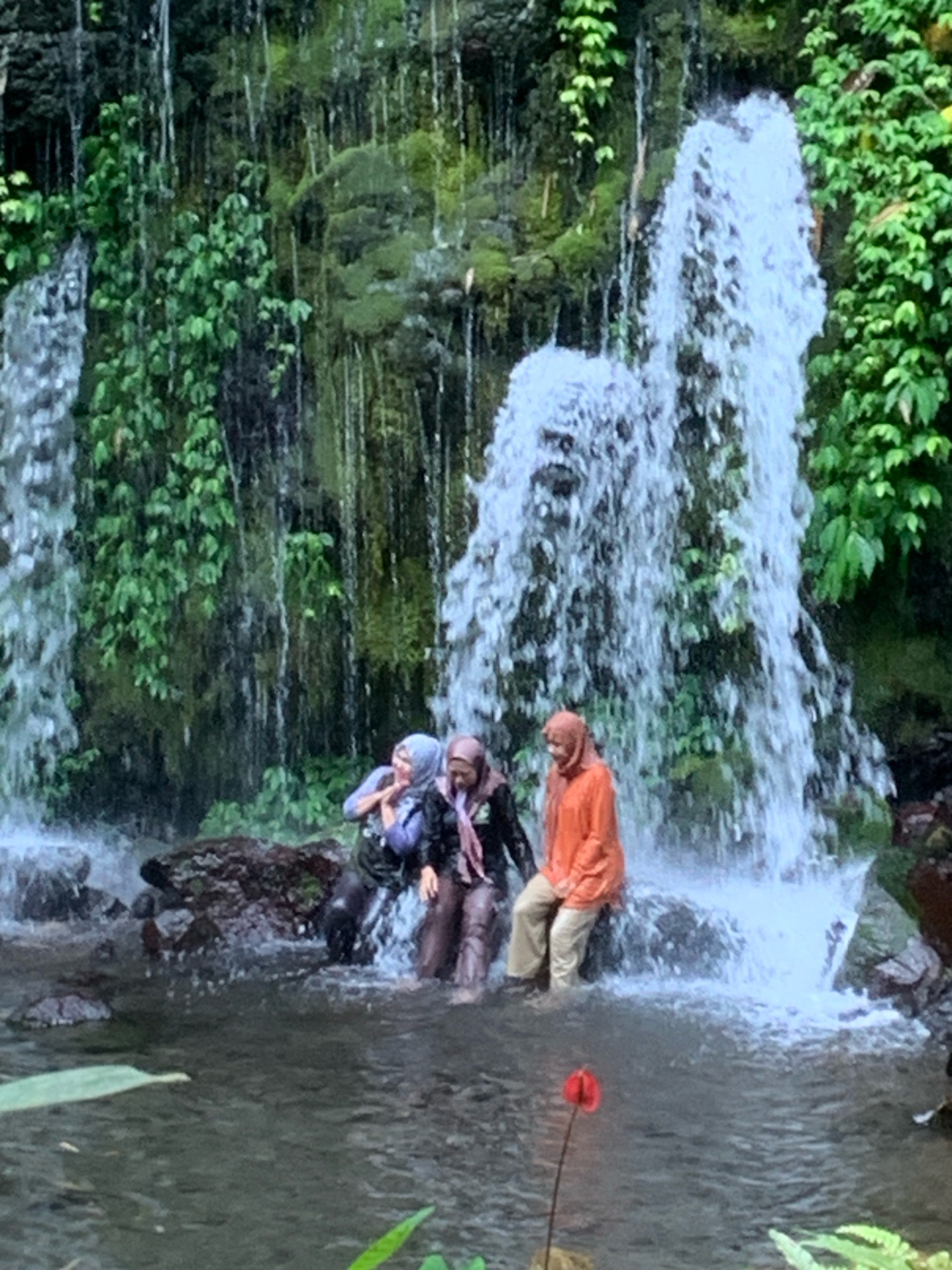 Three individuals, two women and one person whose gender is not discernible, are positioned in the shallow water at the base of a waterfall in Pronojiwo, Indonesia. The two women on the left are wearing hijabs, one in purple and the other in a darker color with a pink scarf. They are partially submerged in the water, with the woman in purple appearing to be speaking to the other. The third individual, wearing an orange headscarf and light-colored pants, is seated on a rock to the right, facing towards the waterfall. The waterfall itself is a significant feature, with water cascading down a moss-covered rock face surrounded by lush green foliage. The water creates a misty spray at the base where the individuals are located. The foreground shows the surface of the water, with a single red flower on a thin stem emerging from it. The overall scene depicts a natural recreational setting at the waterfall.