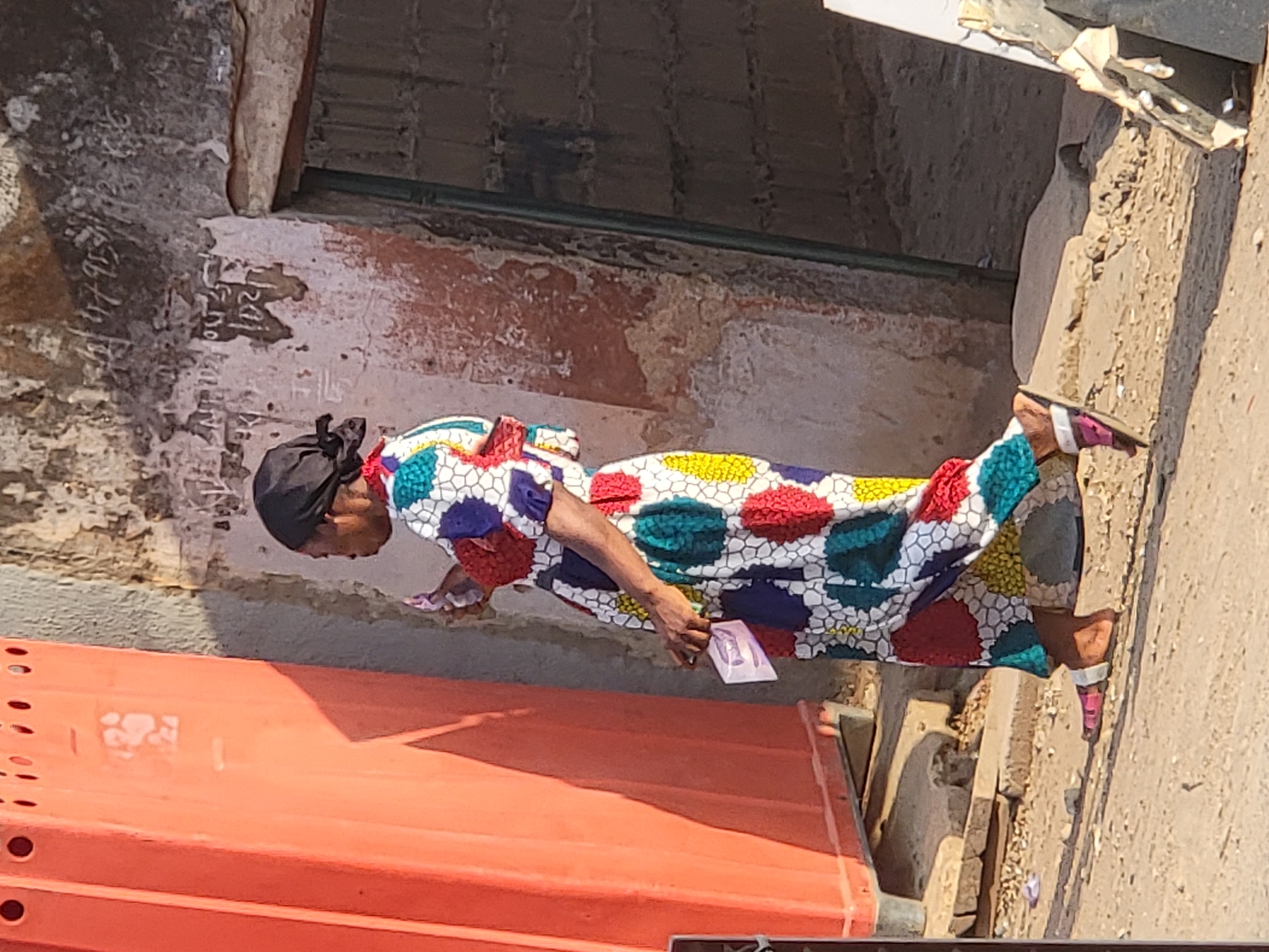 A woman wearing a colorful dress and black head covering stands next to an orange object, possibly examining money in her hands. The location appears to be outdoors, possibly in the city of Maiburiji, Nigeria, based on the prompt's context. The surrounding environment includes walls with visible wear and markings.