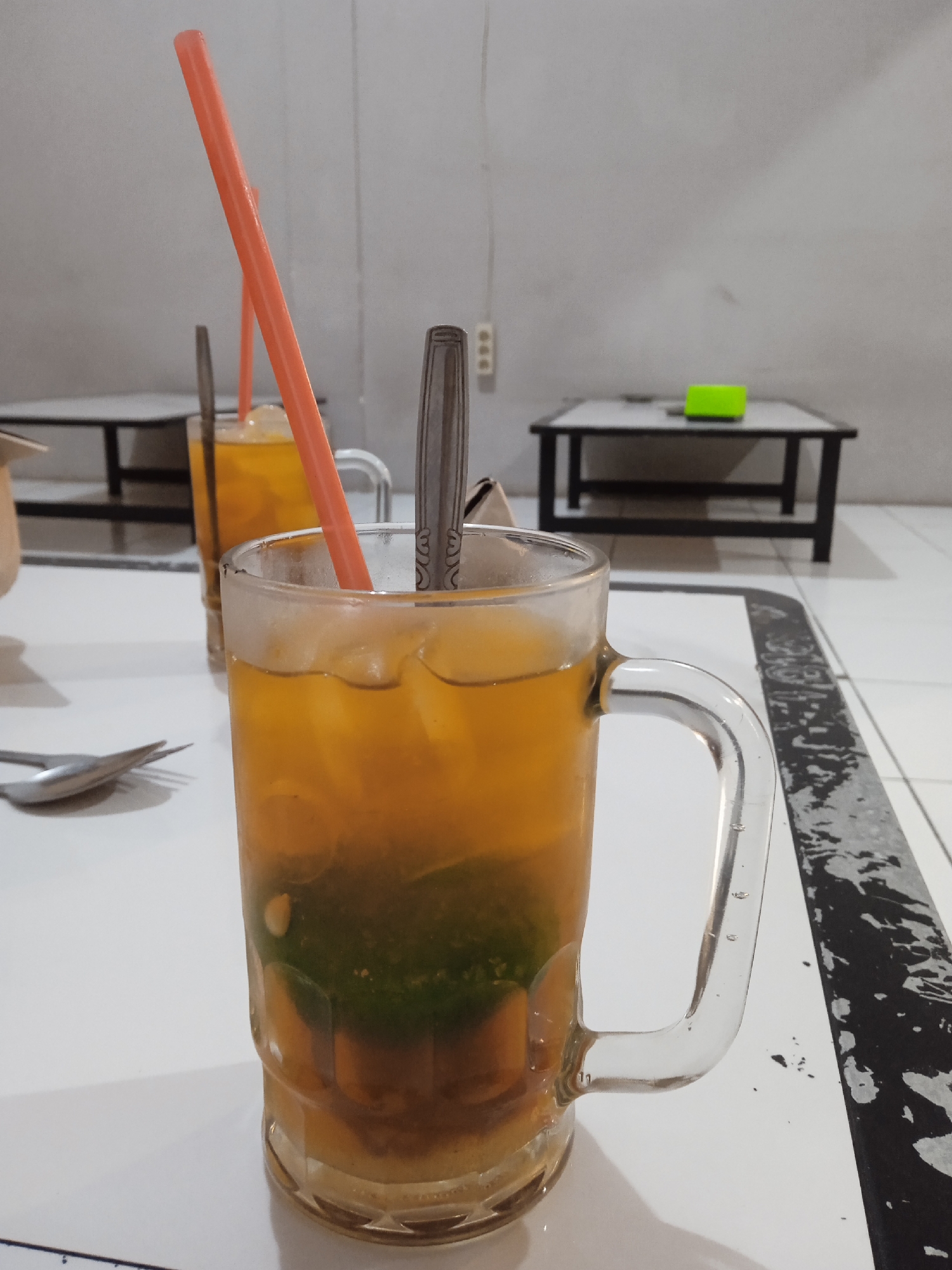 The image shows two clear glass mugs filled with a beverage, likely iced tea or a similar drink, on a white table. A spoon and an orange plastic straw are visible in the foreground mug. The mug in the foreground contains a green ingredient at the bottom. A second similar mug is in the background, also with a straw and spoon. In the background are two small black tables with a white top. The image appears to be taken indoors, with a white wall behind the tables. There is an electrical outlet on the wall behind the tables, and a green object on the table further away. The table is white with black and white decoration on the edge. Given the location, it's plausible this is in Cangkring, Indonesia. No people are visible in the image.
