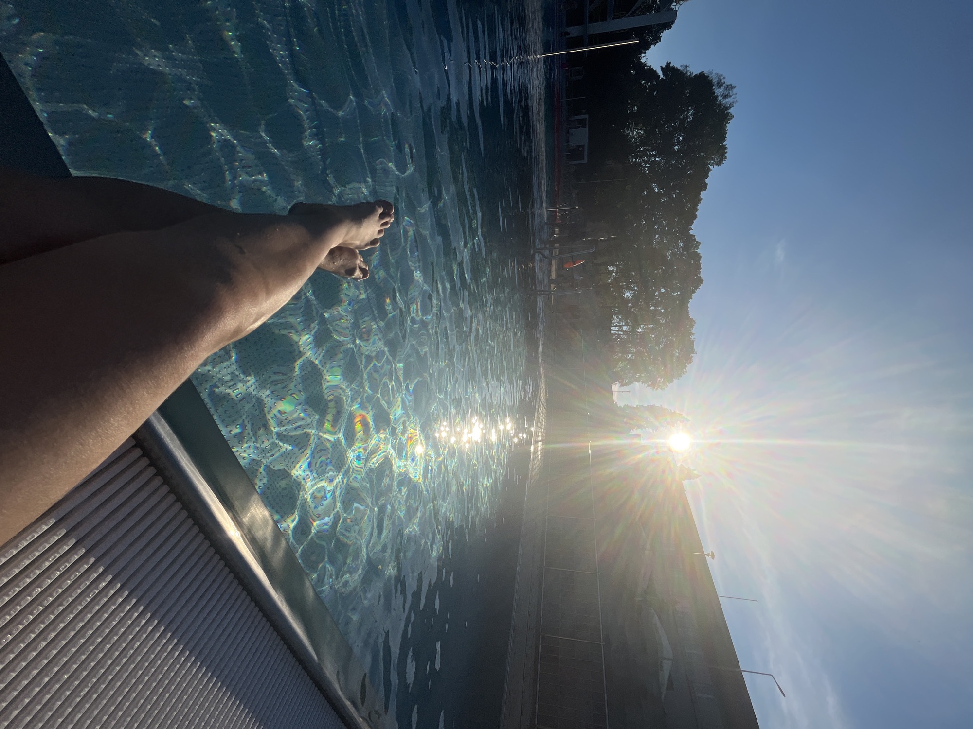 The image depicts a person's legs and feet partially submerged in a swimming pool, with the sun's rays creating a lens flare effect across the water. The pool is surrounded by a tiled edge and a fence. Trees and a building are visible in the background under a clear blue sky. The perspective is from the viewpoint of someone sitting at the edge of the pool.