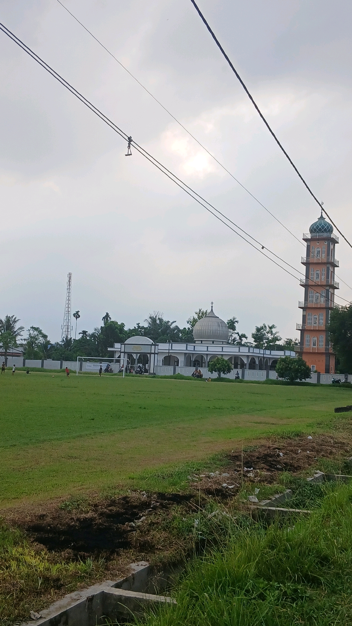 The image depicts a scene in Sunggal, Indonesia, featuring a mosque with an adjacent minaret, set against a cloudy sky. In the foreground, a grassy field is visible, with a few individuals engaged in a game resembling soccer. A telecommunications tower stands to the left of the mosque. The mosque itself has a prominent dome and is surrounded by a white fence. The minaret is tall and orange, with multiple levels and windows. Power lines stretch across the upper portion of the image, with a figure appearing to be walking or perched on them in the distance. The overall environment suggests a public space with recreational and religious facilities.