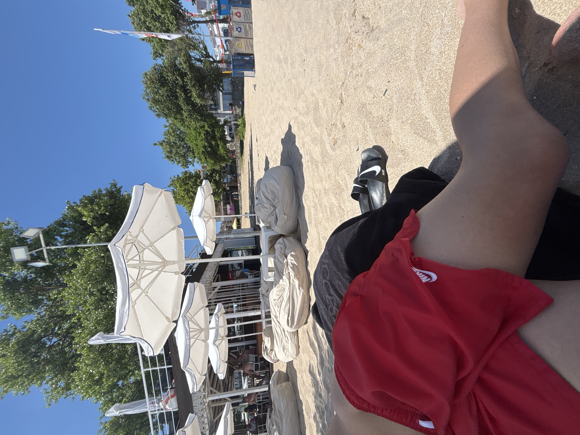 The media file shows a person relaxing on a sandy beach on a sunny day. The view is from a first-person perspective, looking down at the sand and the person's legs. The person is wearing red shorts with a white Nike logo and black slide sandals with white Nike logos. In the background, there are several white beach umbrellas, trees, and a building that appears to be a cafe or restaurant. The sky is clear and blue, indicating fair weather. The scene suggests a leisurely vacation or a day spent enjoying the beach. Some text is visible on the shorts and the sandals. The location is identified as Sozopol, Bulgaria.