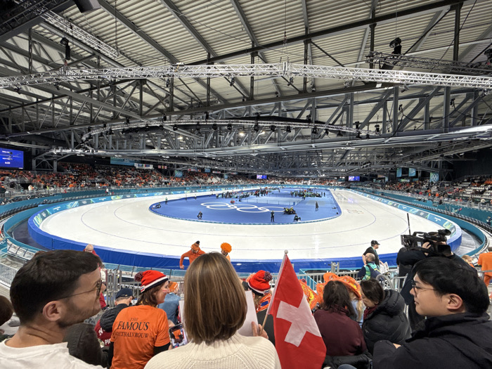 This image presents a wide-angle view of a large, brightly lit indoor speed skating arena located in Rho, Italy. The central focus is an oval-shaped ice track, delineated by a blue barrier. Inside the track, a blue infield area displays the five interlocking Olympic rings on its surface. Numerous individuals, identified as staff or athletes, are dispersed across both the ice track and the blue infield, some appearing to be standing, others potentially kneeling or moving equipment.
Above the track, the venue's ceiling features an intricate metal truss structure supporting an array of lighting fixtures and audio speakers. Tiered spectator seating, largely occupied by a crowd, encircles the entire ice track. Many spectators are observed wearing orange clothing, indicative of support for the Netherlands. Large video screens are visible on the far walls, with one displaying "0:00" and "NL" text.
In the immediate foreground, the backs of several spectators are prominently featured. A person on the left wears a white long-sleeved shirt and eyeglasses, looking towards the ice. To their right, a spectator in an orange t-shirt emblazoned with "THE FAMOUS VOETBALBOUW" wears a red and white striped winter hat. Adjacent to them, a person with brown hair holds a small Swiss flag. Further to the right, a spectator in a dark jacket is visible, with a professional broadcast camera operated by a crew member positioned between the spectator and the rink, suggesting active recording of the event.