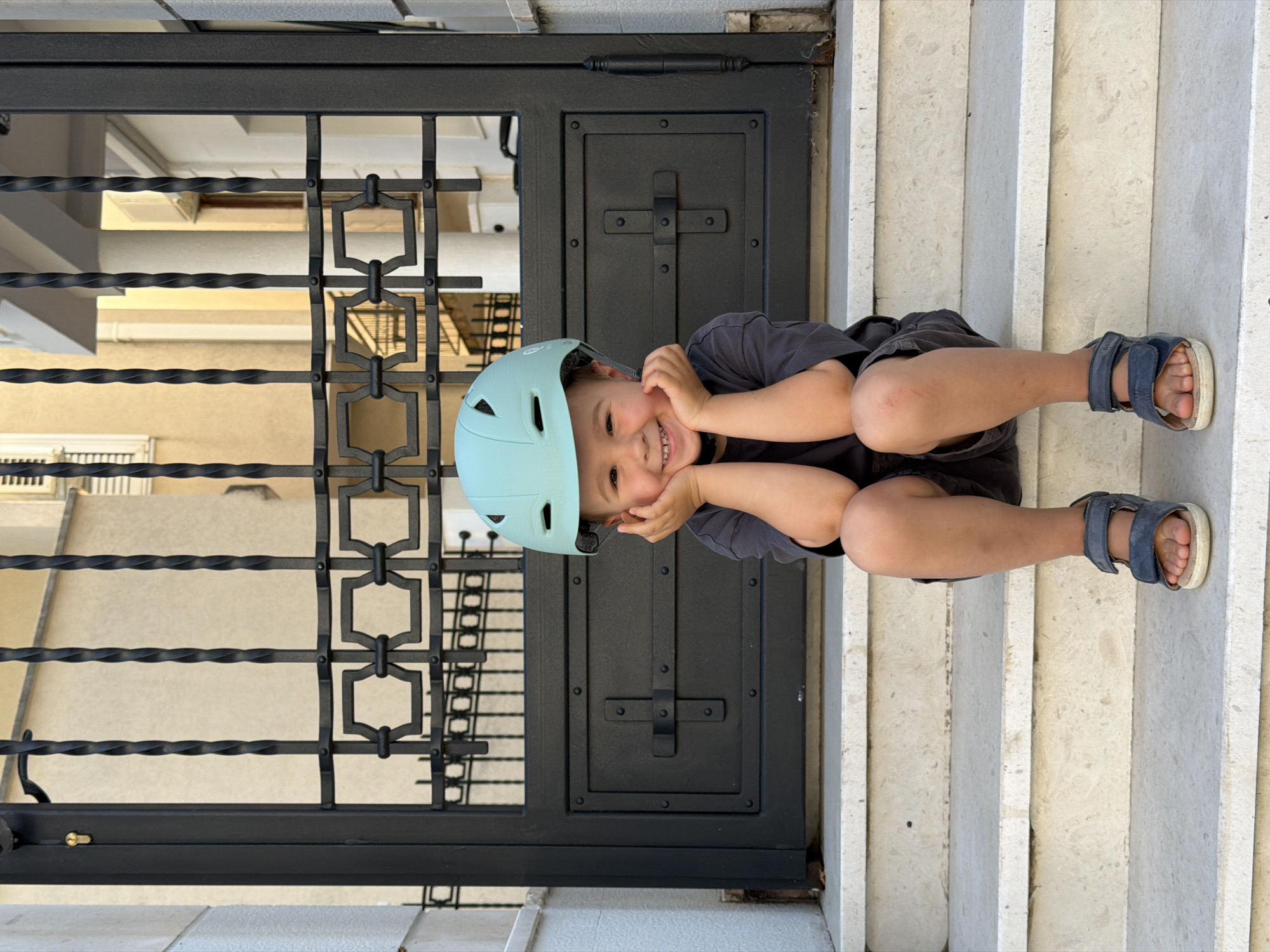 A child wearing a light blue helmet sits on the steps in front of a black metal door, smiling with his hands on his cheeks. The steps appear to be made of a light-colored stone. The child is wearing a gray t-shirt and dark shorts, and blue sandals. The location is Budva, Montenegro, inferred from architectural style and geographical context clues.