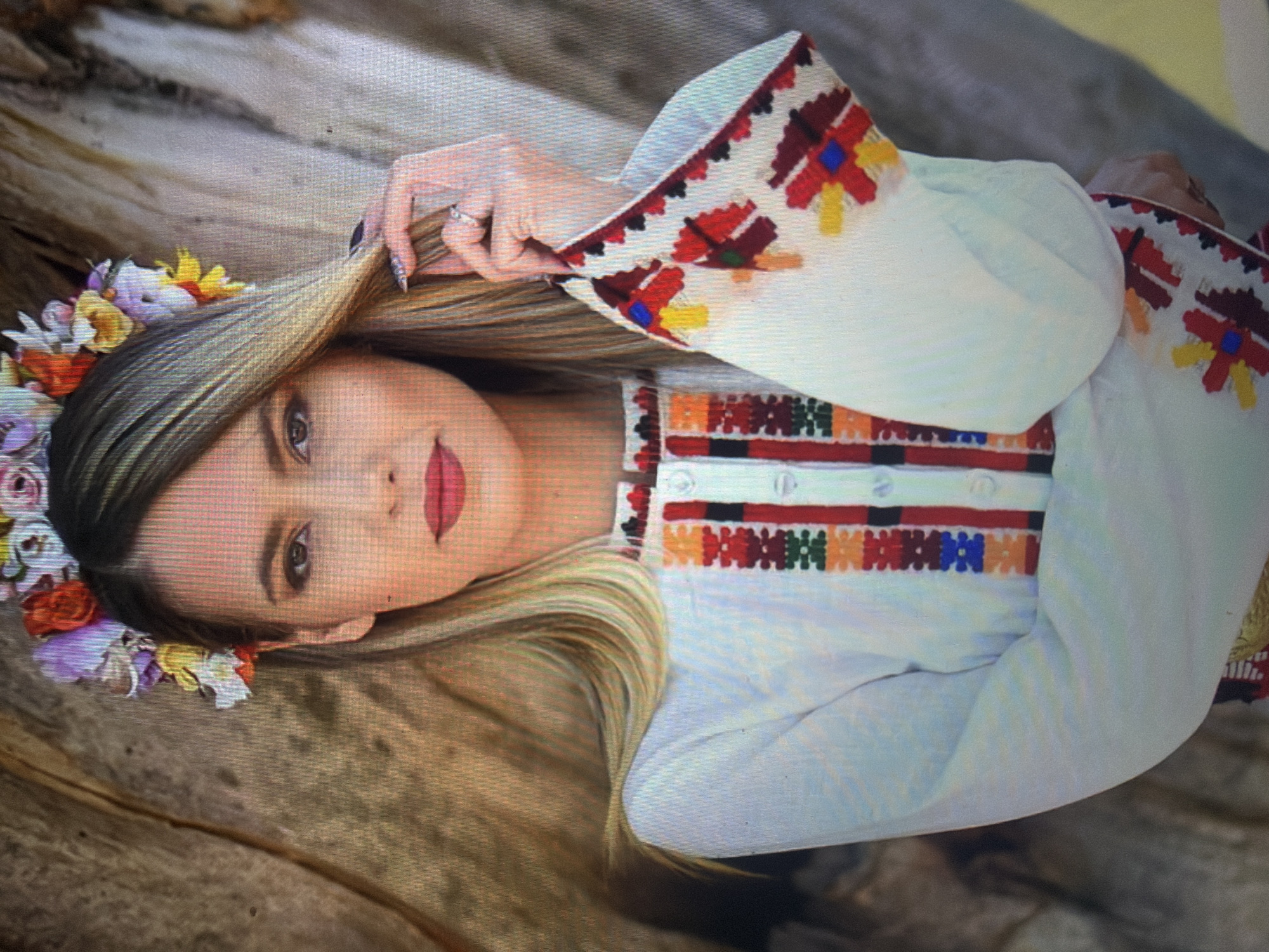 A young adult female is depicted from the waist up, centered against a natural, textured background resembling tree bark. She has long, straight, blonde hair with darker roots, worn down, and is adorned with a floral crown composed of multi-colored flowers including red, orange, yellow, purple, and white blossoms. Her makeup includes dark eyeliner and red lipstick. She is wearing a white long-sleeved blouse featuring intricate, multi-colored traditional embroidery. The embroidery on the central placket consists of vertical bands with geometric patterns in red, orange, green, blue, and black. The wide sleeves display larger, multi-pointed star or cross motifs in red, yellow, and blue, framed by smaller patterned borders. Her left hand is raised to her hair, gently holding a strand near her face; a silver-toned ring with a clear stone is visible on her left ring finger, and her fingernails are painted dark. The scene is located in the city of Shumen, Bulgaria.