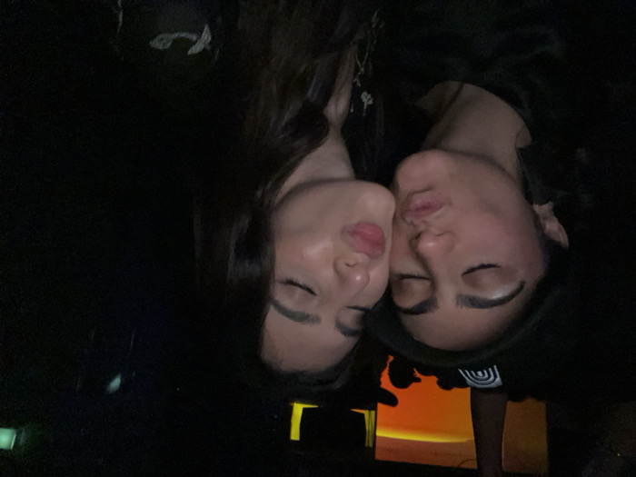 Two women with dark hair and closed eyes are cheek-to-cheek. One is on the left, and the other is on the right. Their lips are pursed. The background is very dark. A screen with an orange glow and black frame is visible at the bottom. The image appears to be rotated.