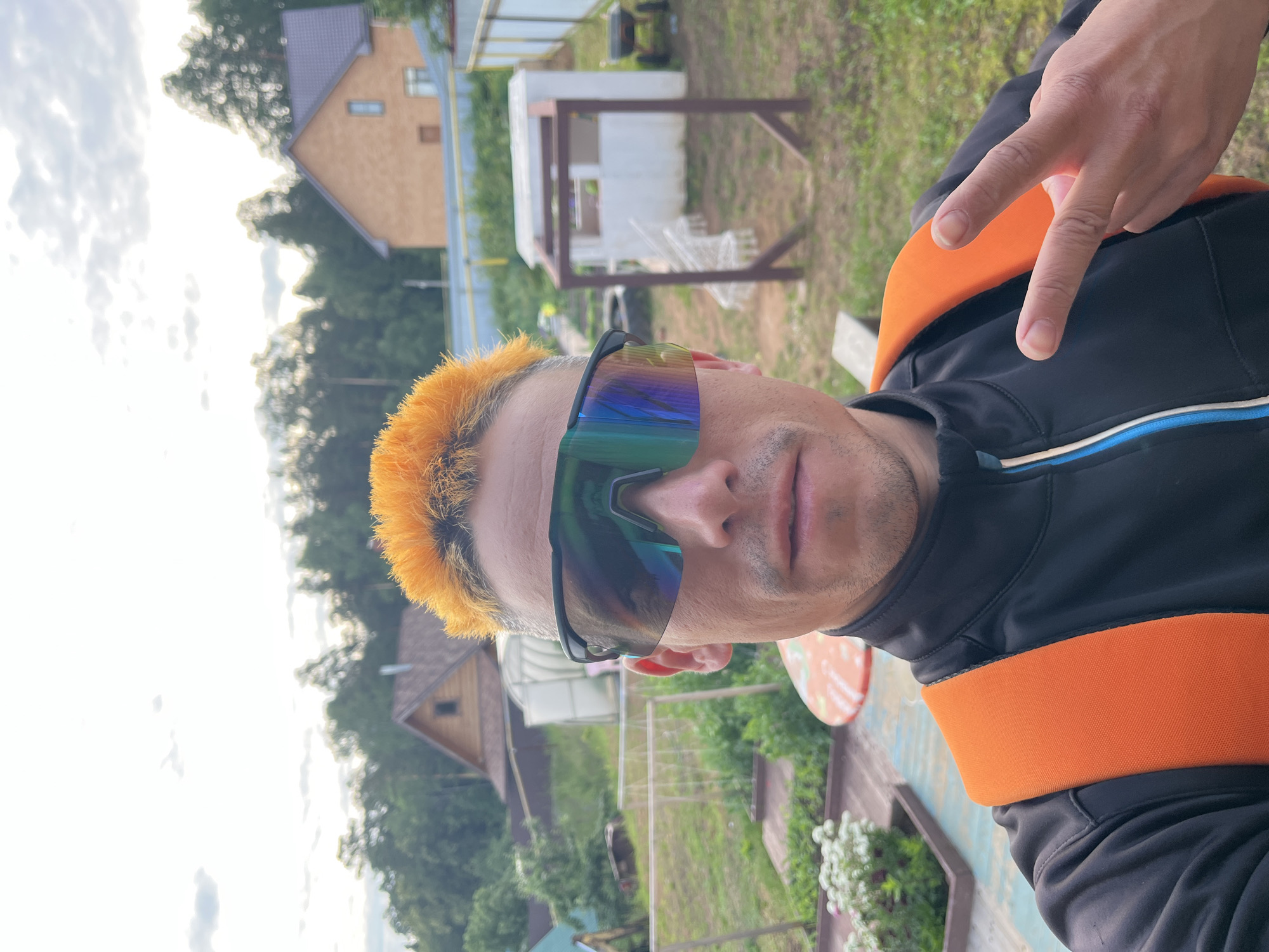 The image shows a man with spiky, bright orange hair and mirrored sunglasses taking a selfie outdoors. He is wearing a black jacket with blue and white accents and an orange backpack strap visible over his shoulder. His right hand is raised, making a peace sign. The background reveals a rural or suburban setting with a wooden house, a greenhouse, a fence, and trees. The sky is cloudy. The scene appears to be in Chaikovsky, Russia, suggested by the architecture and natural environment.