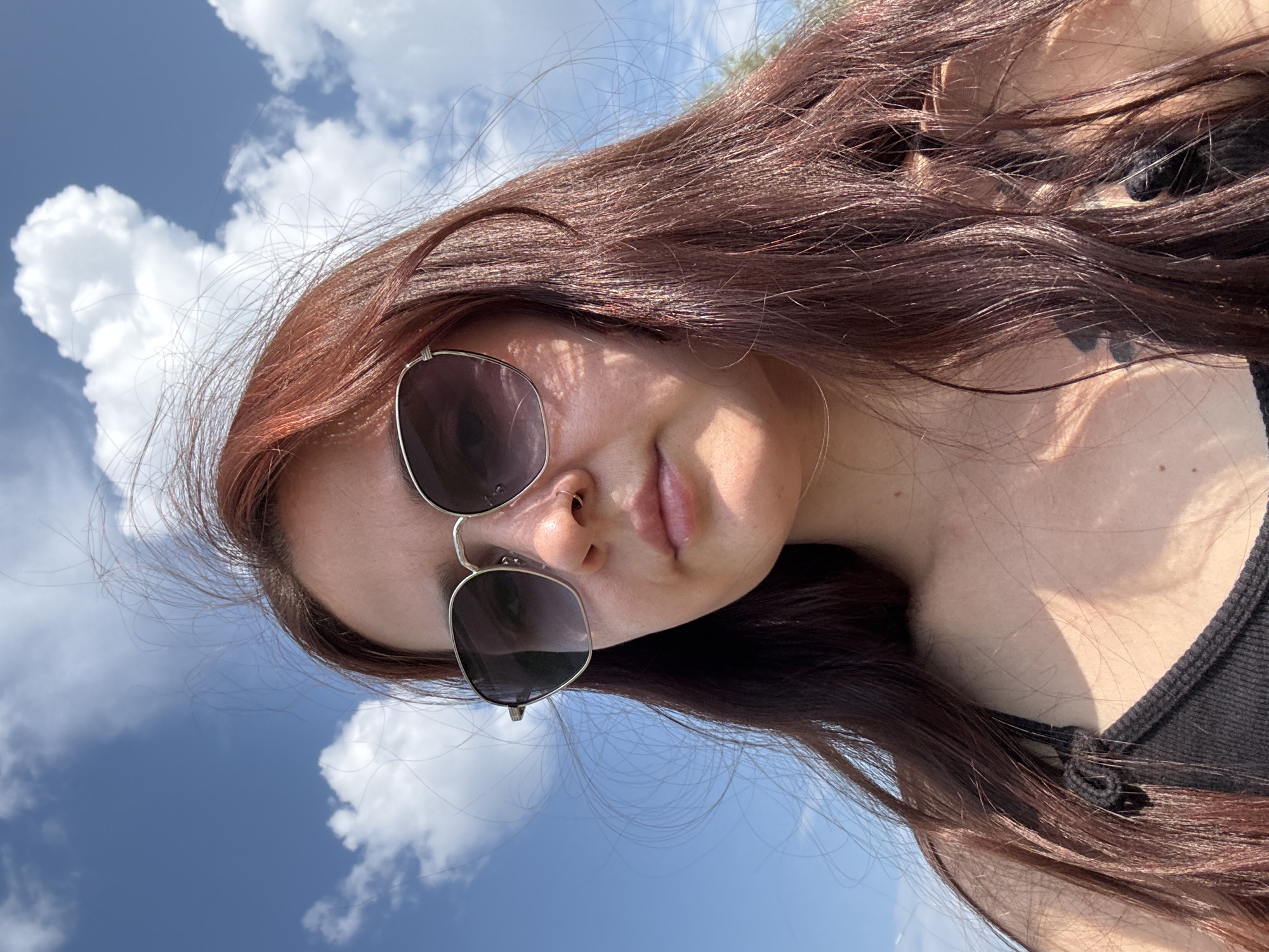 An outdoor, close-up selfie shows a young woman with long, reddish-brown hair and sunglasses against a bright blue sky with scattered white clouds. She is wearing a dark tank top. Sunlight casts shadows on her face and décolletage. The location is unspecified.