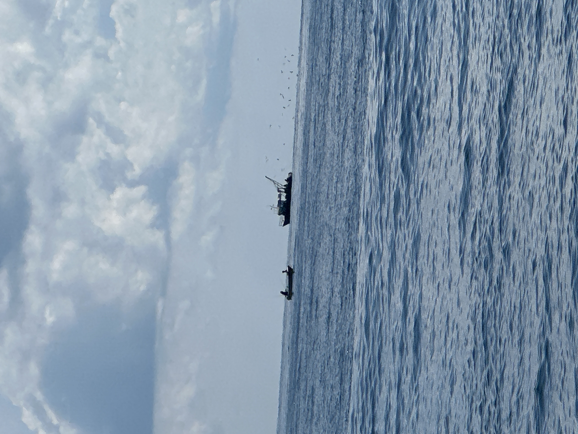 Two small boats and a larger vessel are visible on a body of water under a cloudy sky. The foreground shows the surface of the water with gentle waves. In the mid-ground, to the left, a small dinghy with at least one person aboard is moving across the water. Further to the right, a larger fishing vessel with a crane-like structure is present, and a flock of birds appears to be flying around it. The background is dominated by a sky filled with a mix of dark and light clouds.