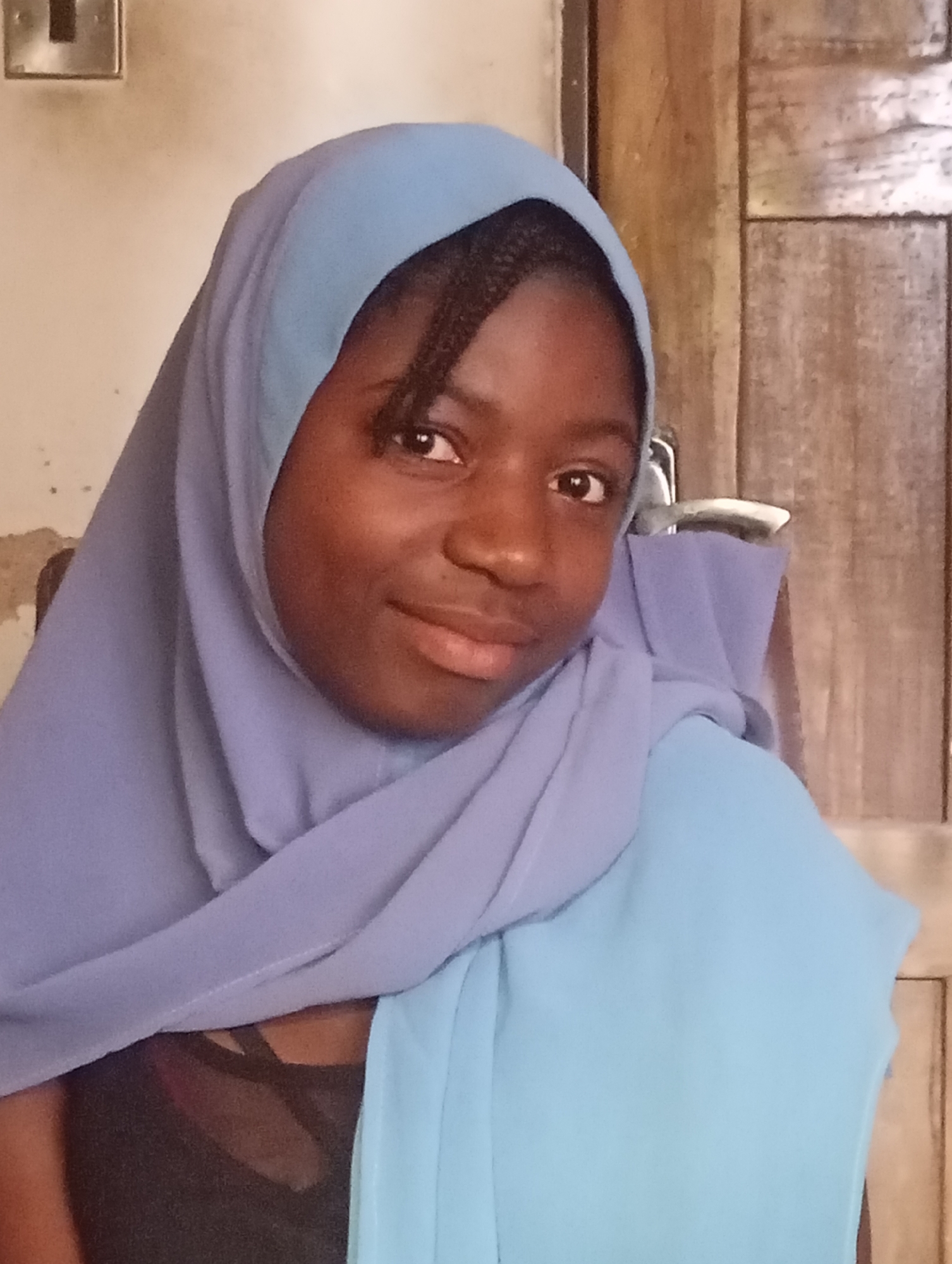 A young Black woman is centered in the frame, wearing a blue hijab. Her dark hair is visible in braids along her forehead. She has a warm, subtle smile, and her gaze is directed towards the viewer. Below the hijab, a black top is visible. The background is a neutral wall to the left, with a light switch and a wooden door to the right. The location is Unknown, Unknown.