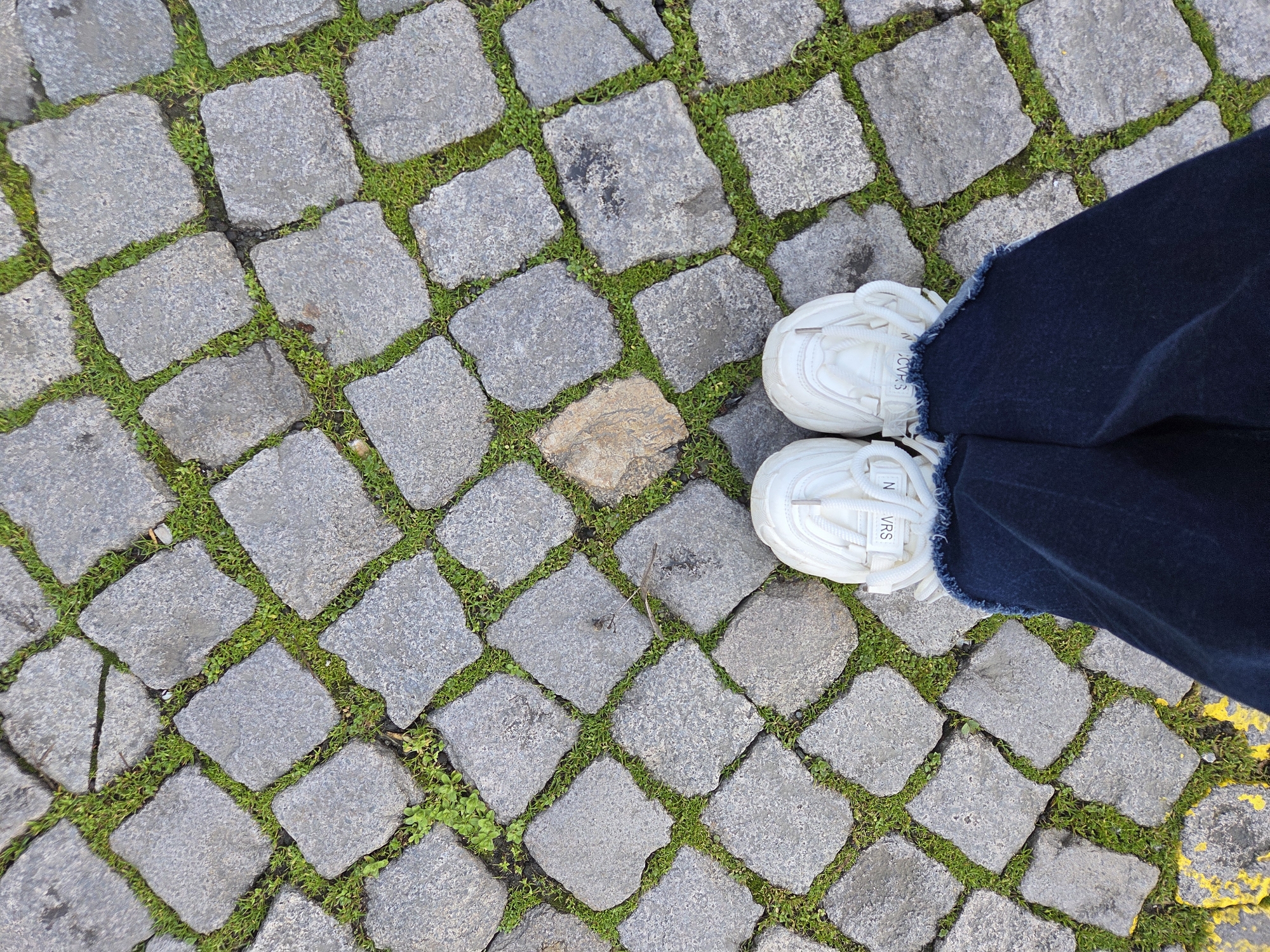 A top-down perspective captures the lower extremities of a person standing on cobblestone pavement in Burgas, Bulgaria. The subject wears dark blue denim pants with frayed cuffs and white athletic shoes with chunky soles, white laces, and visible white fabric straps featuring inverted black lettering ("S V R N"). The pavement consists of irregular grey cobblestones, with vibrant green grass and weeds growing between the gaps. A light brown, irregularly shaped stone or piece of debris is present in the upper-middle section of the cobblestones. In the lower-left corner, a yellow painted line is visible along the edge of the pavement. No actions or interactions are depicted beyond the static presence of the person's feet.