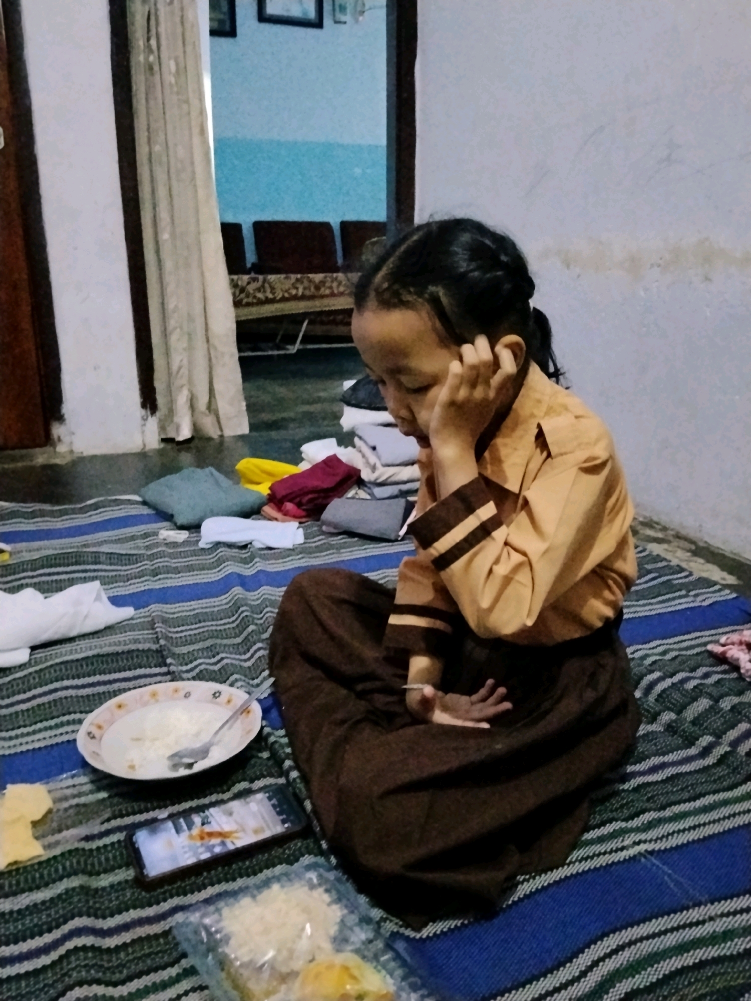 A young girl, likely Indonesian given the location context of Kejawan, is seated on a striped mat. She wears a tan school uniform with dark brown stripes on the sleeves and collar, and a dark brown skirt. Her hair is styled in braids. Her right hand is pressed against her ear, and she appears to be looking down with a pensive or tired expression.
In front of her on the mat are several objects. To her left, a white plate with a spoon contains a small amount of white food, possibly rice. Next to the plate is a smartphone displaying an image. Further to the right, a clear plastic container holds more white food and a yellow pastry. A piece of yellow food, perhaps cheese or a snack, is also visible on the mat. To her left, a pile of folded clothes is present.
The setting appears to be indoors, with a wall in the background and part of a doorway with a curtain visible on the left. The lighting suggests it might be evening or indoors with artificial light, as there is no indication of natural daylight. The overall scene conveys a sense of quiet or perhaps a moment of rest after a meal. There is no visible text in the image.