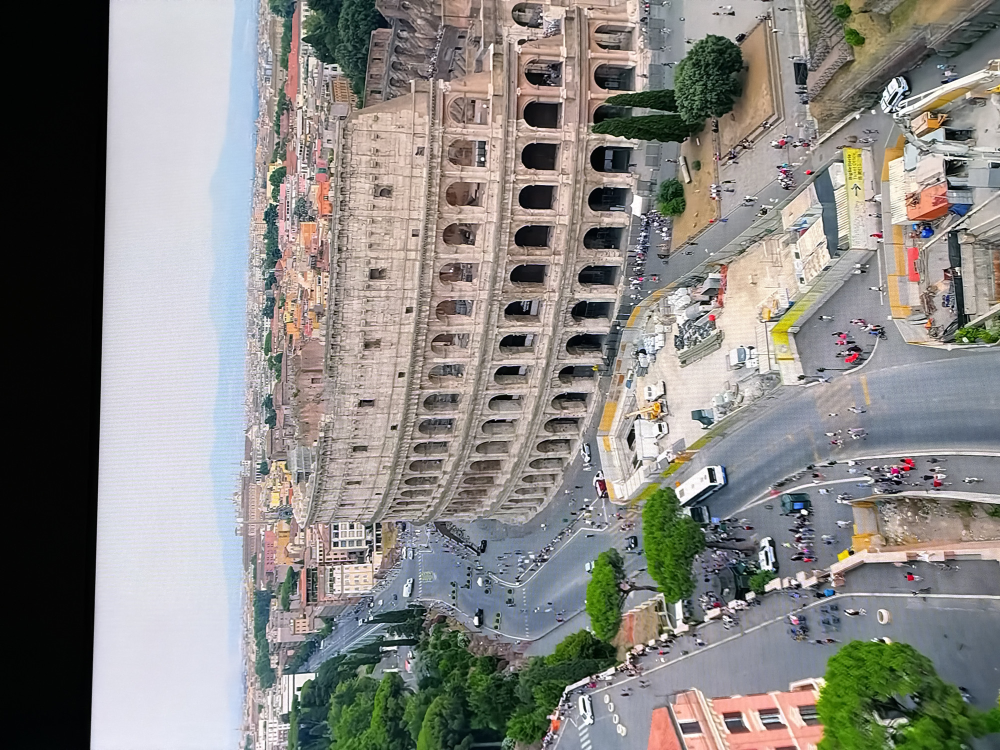 The image depicts an aerial view of the Colosseum in Rome, Italy, with a cityscape extending into the background. Numerous tourists are visible walking around the Colosseum and along the adjacent road. The area around the Colosseum shows signs of ongoing construction or renovation, with barriers, scaffolding, and construction materials present. Several vehicles, including buses and cars, are on the road. The sky is overcast, and distant mountains are visible on the horizon. The image does not show any elements related to Chirchik, Uzbekistan.