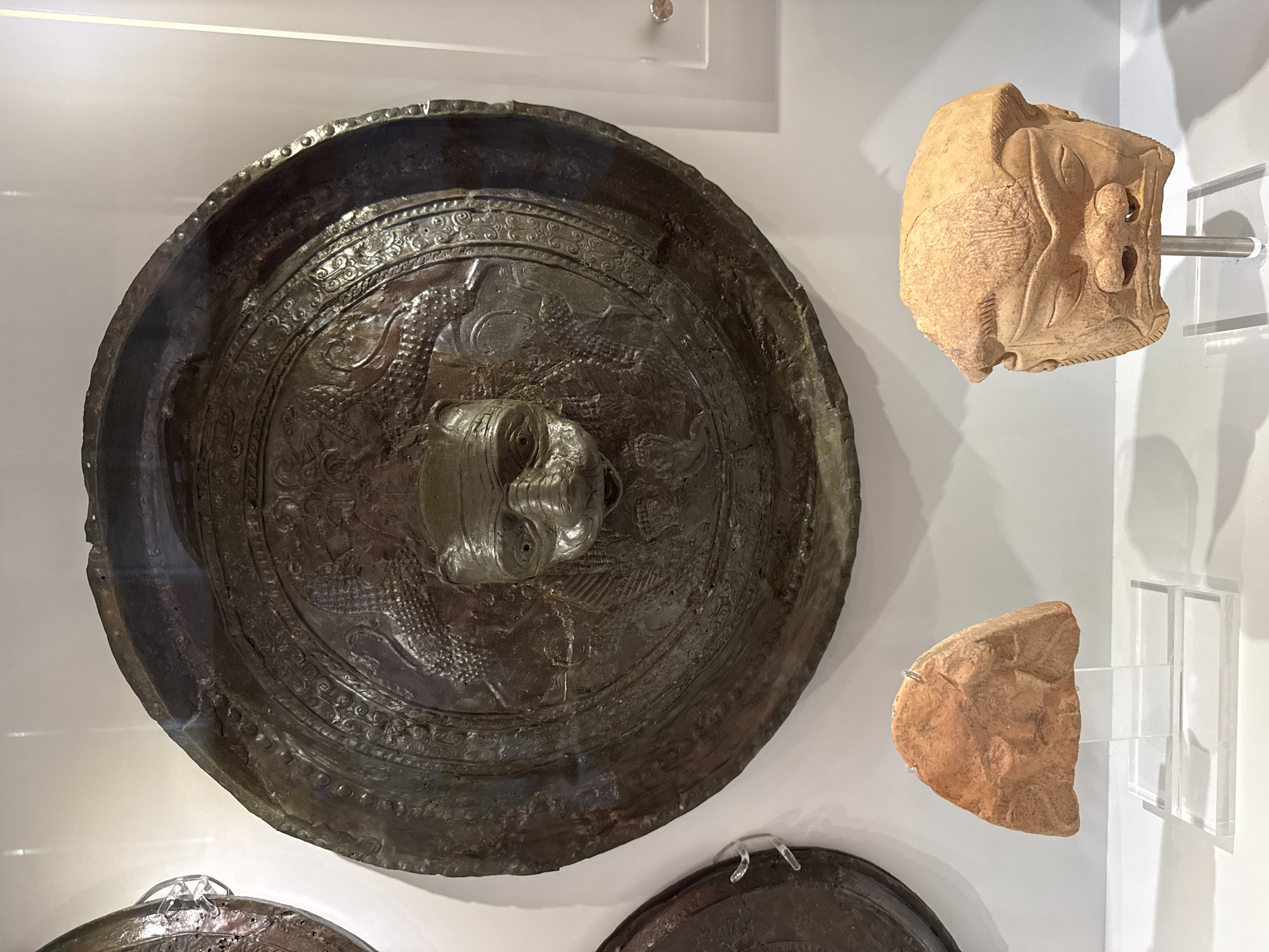 The image depicts an archaeological display within a museum, likely the Heraklion Archaeological Museum in Heraklion, Greece, given the nature of the artifacts.
The central and largest object is a circular, dark metallic shield, possibly bronze, with a prominent, raised, grotesque face at its center. The face has wide eyes, a flattened nose, and an open mouth. Surrounding the central face, the shield is intricately decorated with embossed patterns and what appear to be serpentine or draconic figures. The rim of the shield is rough and shows signs of age and wear.
To the lower left of the shield, a small, reddish-brown clay fragment is displayed on a clear acrylic stand. This fragment appears to be part of a larger object, possibly a vessel or sculpture, with faint markings suggesting a face or design.
To the lower right of the shield, a larger, light brown ceramic mask is mounted on a metal rod within a clear acrylic stand. This mask has a stern, somewhat exaggerated facial expression with deep-set eyes, a broad nose, and prominent cheekbones. The mask is detailed with lines suggesting hair or a headdress and some scarring or texture on its surface.
The objects are set against a plain, light-colored wall, indicative of a museum exhibit. The lighting appears to be artificial, illuminating the artifacts clearly. The overall scene conveys a sense of historical significance and the preservation of ancient artifacts.