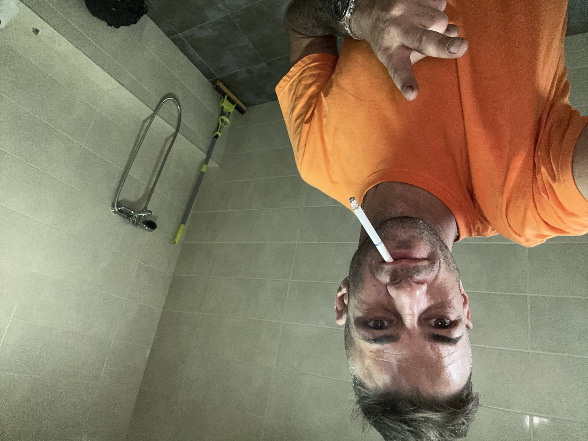 A man with short, spiky hair and a slight stubble is in a bathroom, holding a cigarette in his mouth. He is wearing an orange t-shirt and a watch on his left wrist. His right hand is raised, with his index finger extended. Behind him, on the right side of the frame, is a shower head attached to the tiled wall and a mop with a yellow sponge. The floor is tiled in a dark gray color. The overall environment appears utilitarian and possibly recently cleaned, with beige tiles on the walls and a gray tiled floor. The lighting is somewhat dim. This scene is located in Piperevo, Bulgaria.