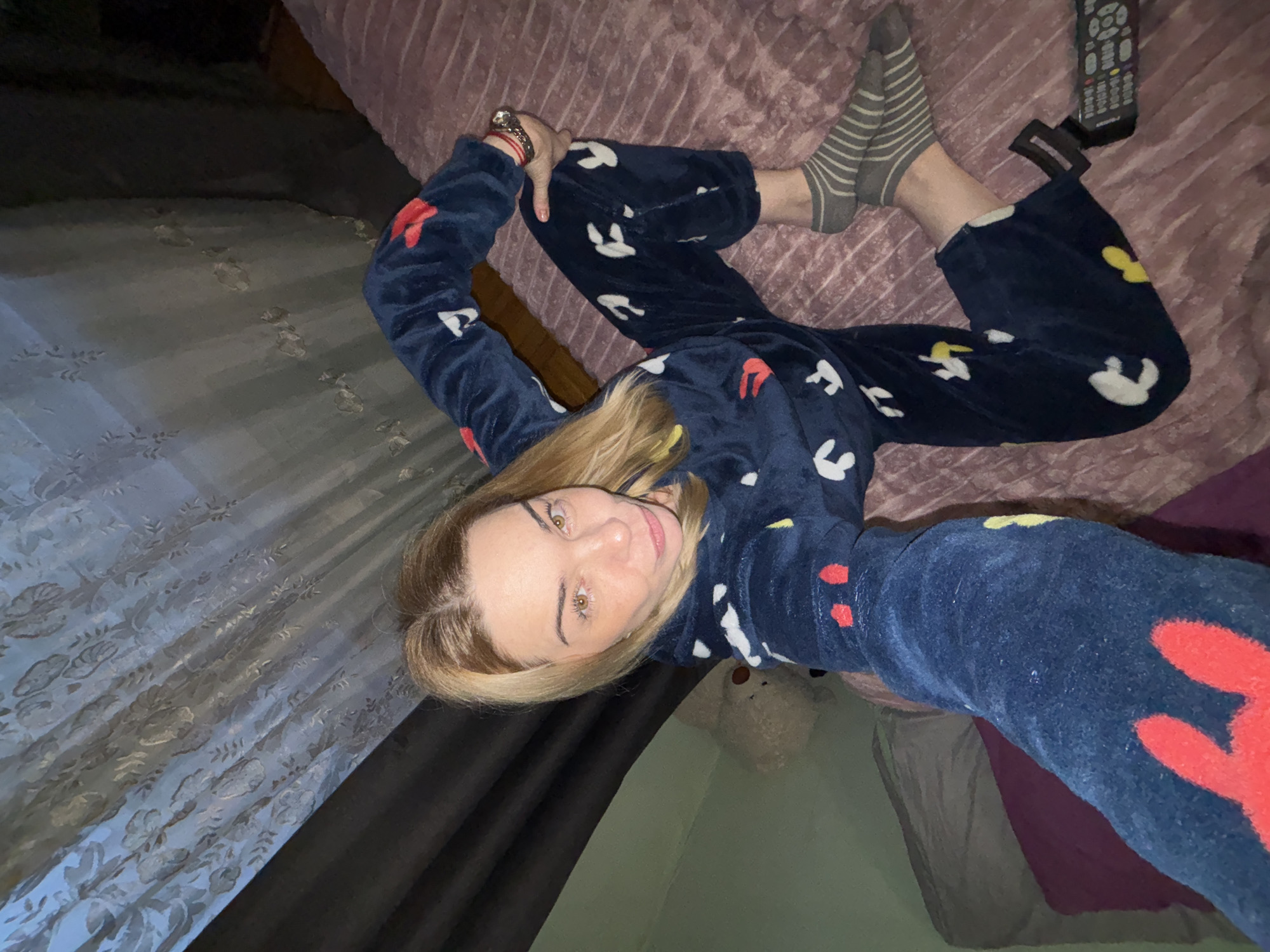 A woman lies on a bed covered with a pink textured blanket and a dark blue patterned pajama set in an indoor setting. She is wearing gray and white striped socks on one foot. A black remote control rests on the bed next to her foot. A curtain hangs to the left of the bed. A teddy bear is on the floor beneath her. The lighting is dim. The city is unknown.