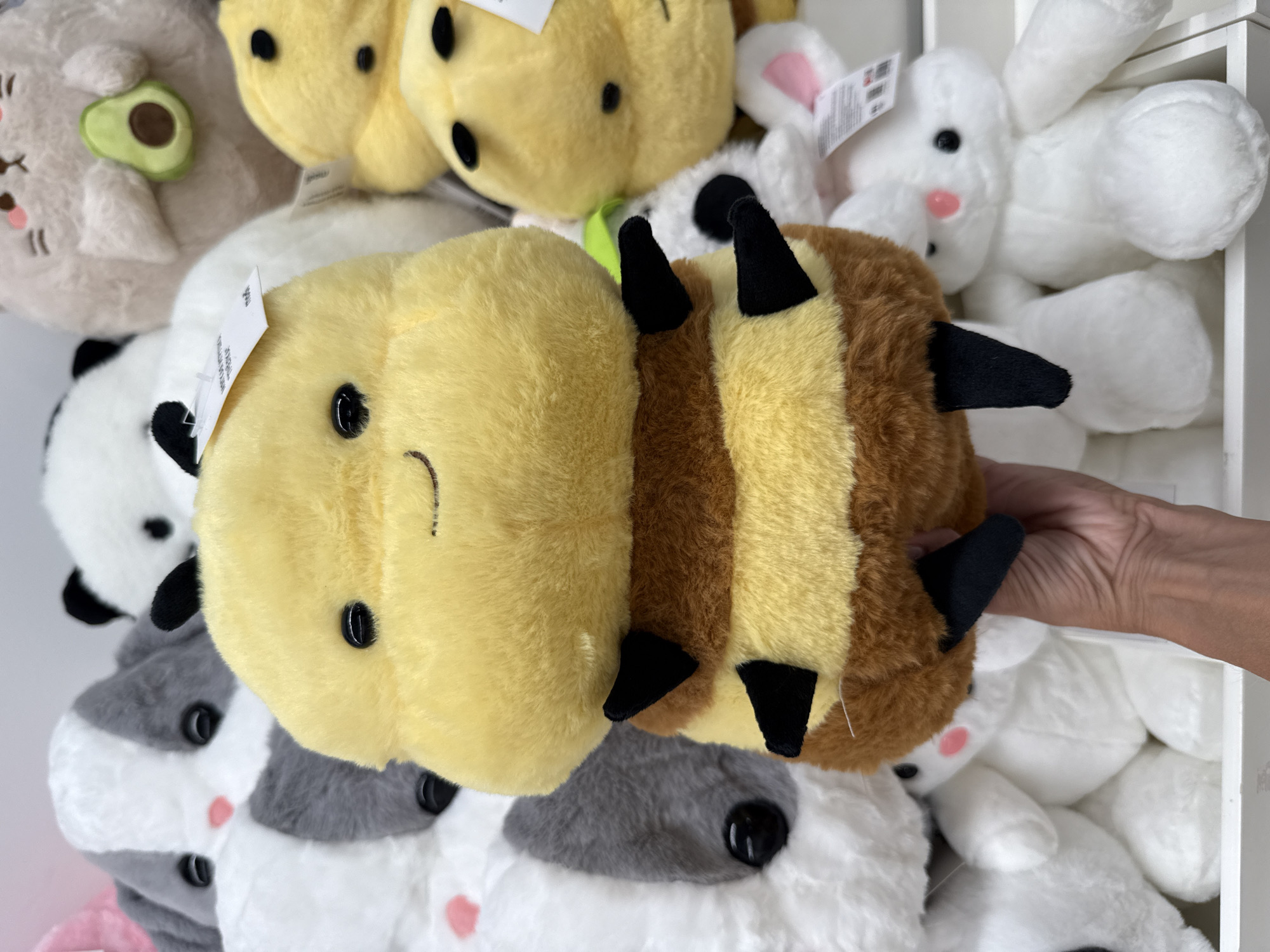 A person's hand is holding a plush bee toy in the foreground. The bee toy is yellow with brown stripes and black legs. Behind the bee toy, there are several other plush toys visible, including a panda, a cat holding an avocado, and a white rabbit. The toys are arranged on shelves in what appears to be a store. The background is out of focus, but it suggests an indoor environment with lighting. The location of Sosnovtsy, Russia is not discernible from the image.