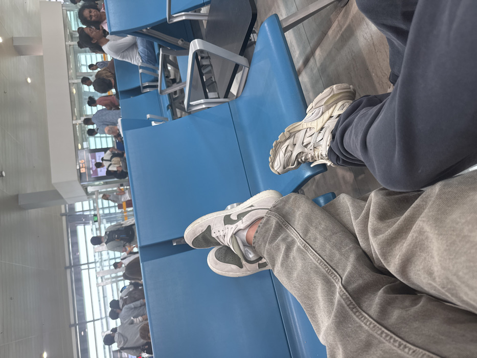 The image depicts an interior scene within an airport waiting area, specifically at a departure gate, likely Abeid Amani Karume International Airport (ZNZ) in Zanzibar, Tanzania.
In the immediate foreground, the lower bodies and feet of two individuals are visible. The person on the left wears light gray, possibly stone-washed, denim pants. Their right foot is elevated, clad in a light gray and dark green athletic shoe (resembling a Nike Jordan 1 Low), and rests on the light blue seat directly in front. The person on the right wears dark blue sweatpants. Their left foot is similarly elevated, wearing a cream-colored, multi-paneled athletic shoe with a prominent sole (resembling a Balenciaga Track Sneaker), also resting on an adjacent light blue seat. Both individuals are positioned as if seated, with their feet elevated onto separate seats.
The environment features multiple rows of light blue, metal-framed airport tandem seating. In the midground and background, various other passengers, appearing diverse in ethnicity and dressed in casual attire, are present. Some are standing near a boarding gate marked "G6," while others are seated further back or moving through the terminal. The area is brightly illuminated by natural light entering through large, rectangular windows that span the background. The ceiling is high and white, featuring recessed lighting. The floor is a light, neutral color. No direct interaction is discernible between the two foreground subjects or with other passengers.