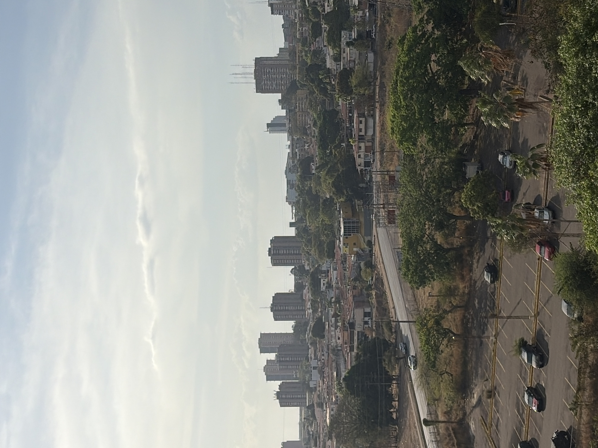 The image shows a cityscape from a high vantage point, likely a building overlooking Ciudad Guayana, Venezuela. The sky is overcast with light-colored clouds. In the distance are several tall apartment buildings densely clustered together. Below, there is a mixture of residential buildings and green spaces composed of trees. A road runs alongside a parking lot with cars parked in designated spots. The road is lined with lampposts.