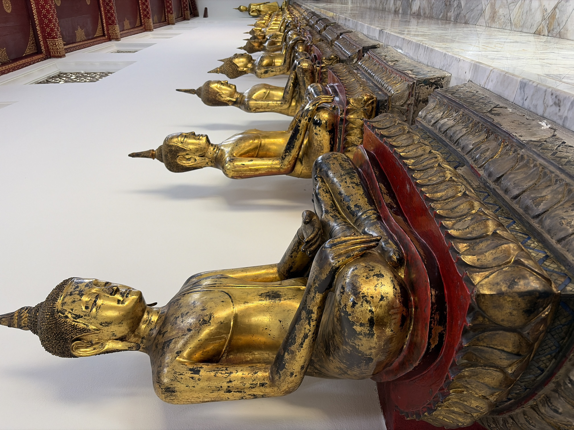This image depicts a row of gilded Buddha statues seated in meditation posture along a white wall inside a temple. The statues are adorned with intricate bases and are illuminated by natural light. The ceiling features ornate red and gold detailing, typical of Thai temple architecture. This scene is characteristic of Wat Pho, one of Bangkok's oldest and most famous temples, known for its reclining Buddha and numerous other Buddha images. The statues are arranged in a line, suggesting a processional or devotional context. The environment is serene and orderly, with a marble floor reflecting the light. There are no people or animals visible in the frame.