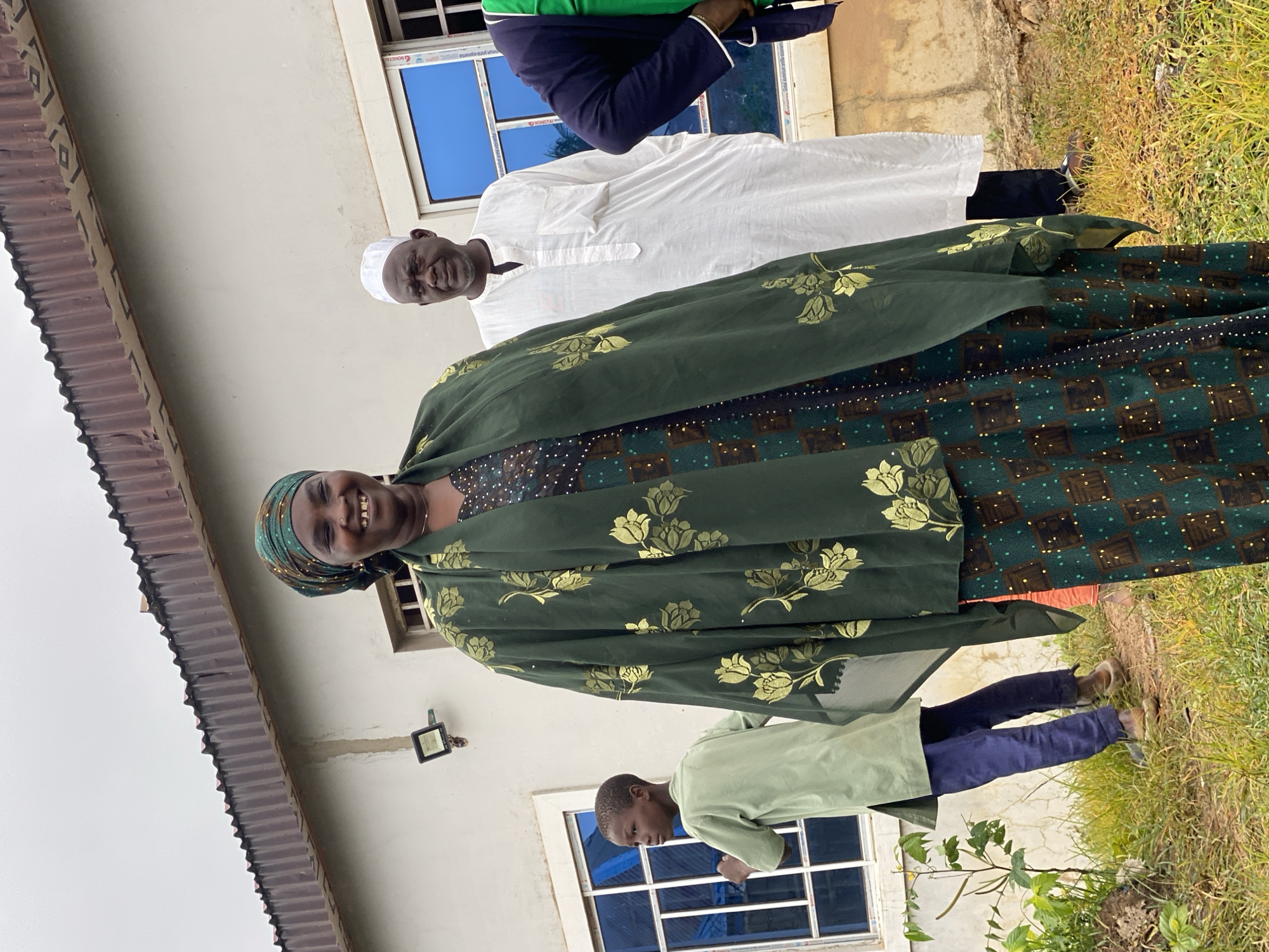 The image shows a group of people in front of a building in Kawo, Nigeria. A woman in a green headscarf and matching green patterned dress is in the foreground, smiling broadly. She is wearing a dark green shawl with gold floral patterns. Behind her, a man in a white cap and white robe stands, looking towards the left. To the left of the woman, a young boy in a light green shirt and dark pants is looking to the left. The building behind them is light-colored with a corrugated metal roof and blue-tinted windows. The ground in front of the building is covered with grass and dirt. The sky is overcast.