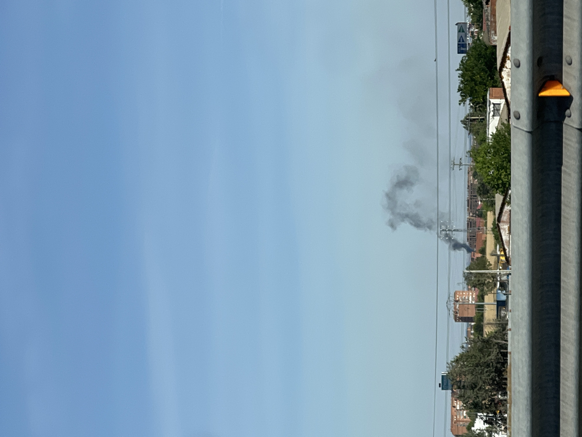 The image shows a hazy, blue sky over a cityscape. In the middle ground, there is a plume of black smoke rising from what appears to be a construction site or industrial area. Several buildings, including some with red brick facades, are visible behind trees and other structures. Power lines crisscross the scene. In the foreground, a grey guardrail with visible rivets and an orange reflector is present. The scene suggests a possible fire or industrial incident in the outskirts of Madrid, Spain.