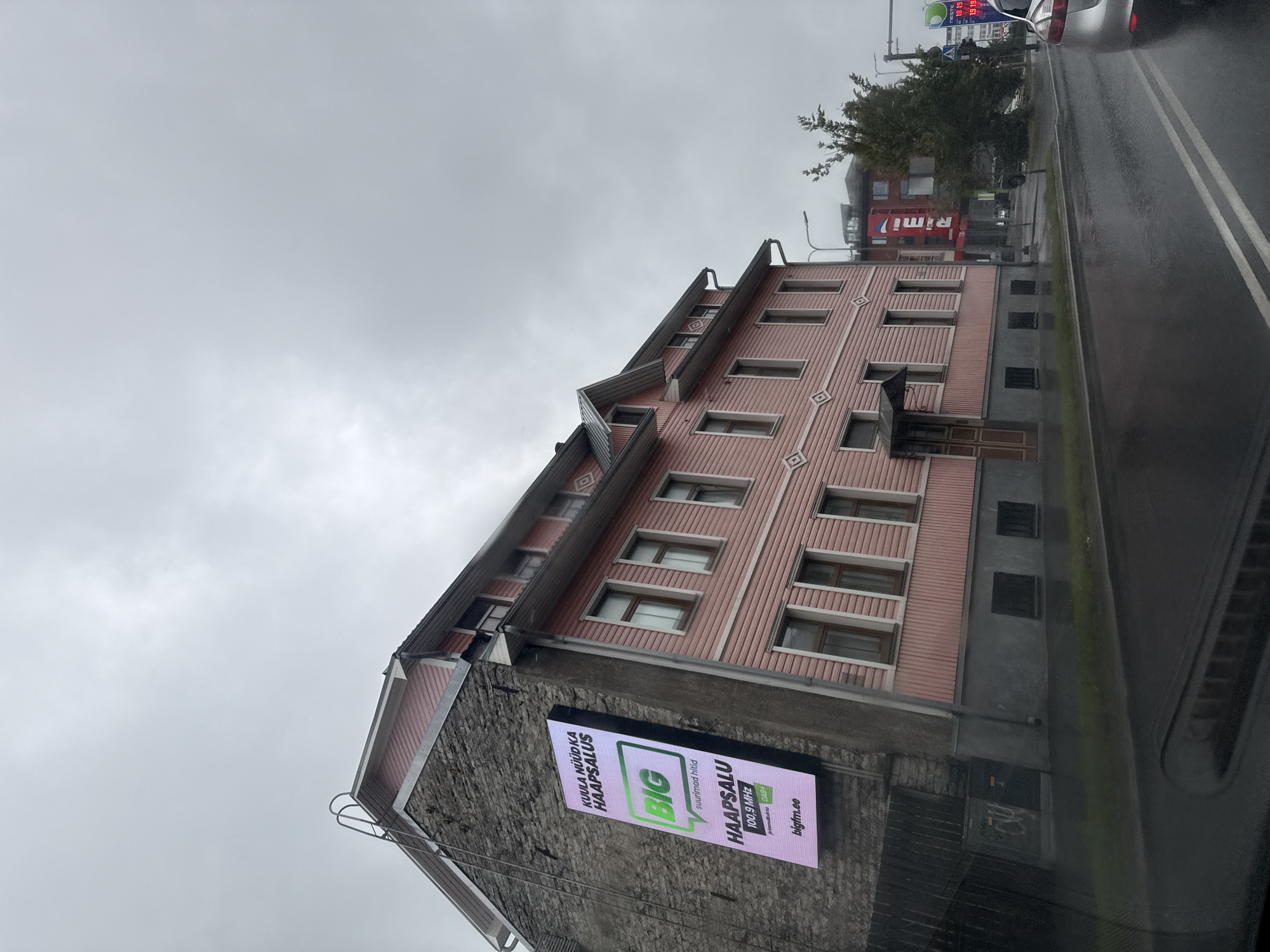 The image captures a street scene in Tallinn, Estonia on a cloudy day. A pink wooden building with a stone facade stands prominently on the left, displaying a large digital advertisement. The advertisement features the text "KUULA NÜÜD HAAPSALUS," "BIG," "suurimad hitid," "HAAPSALU 100.9 MHz," and "bigfm.ee." Adjacent to this building, another pink wooden structure with multiple windows and decorative elements is visible. The street in front is wet, indicating recent rain, and a silver car is parked on the right side of the road. Further down the street, a Rimi supermarket sign is visible, along with trees and streetlights. The sky is overcast, contributing to a muted overall tone of the photograph.