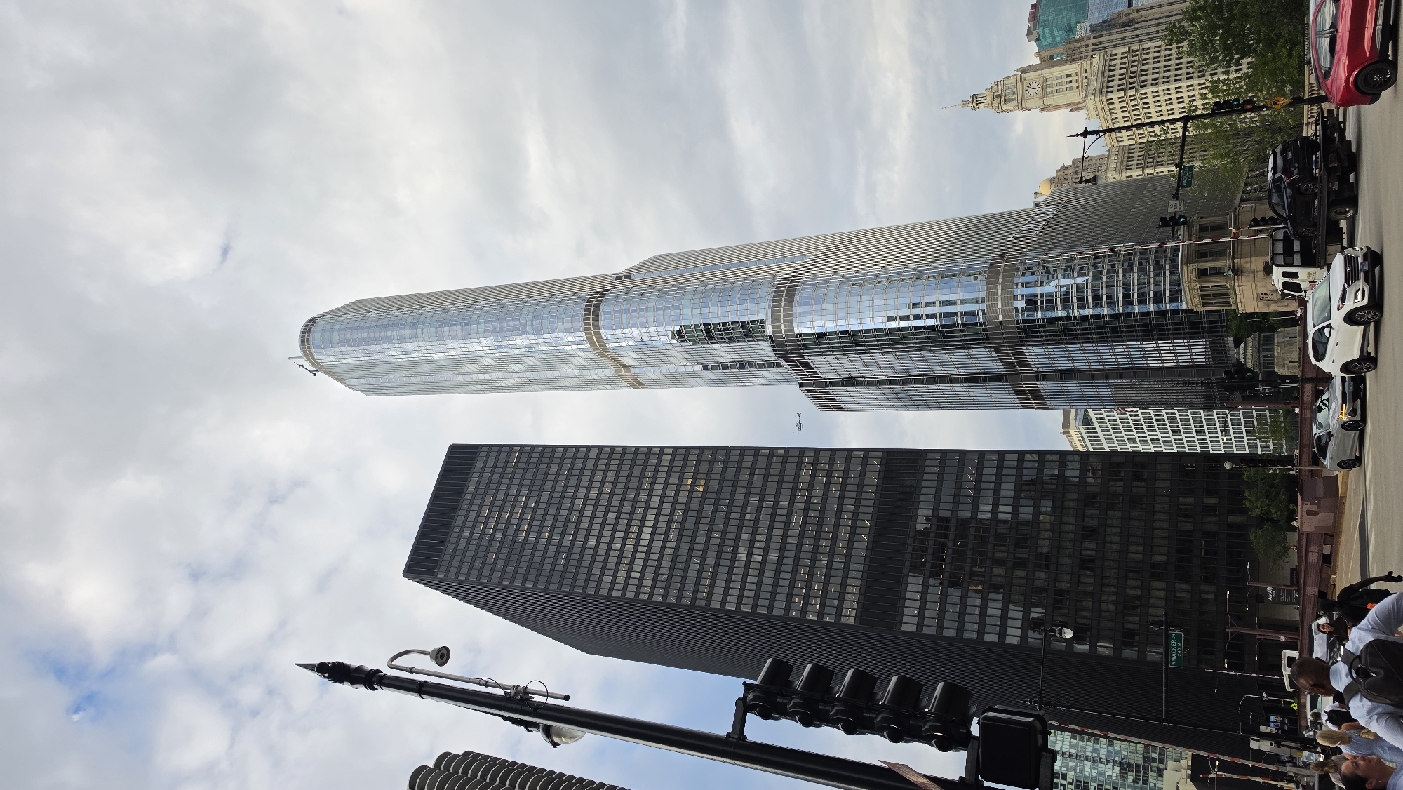 This image, taken from a street-level perspective in Chicago, USA, captures a bustling urban scene dominated by towering skyscrapers. In the foreground, a traffic light with black casing hangs prominently on a pole, with multiple vehicle lanes visible below. Cars, including a white SUV and a silver sedan, are stopped at the intersection. Several pedestrians, including a man with a backpack, are also present.
The background is defined by a striking contrast between architectural styles. To the left stands a dark, rectangular skyscraper with numerous windows. Adjacent to it, and dominating the center of the frame, is the Trump International Hotel and Tower, a sleek, glass-clad skyscraper with a distinctive curved facade that reflects the cloudy sky. Further in the distance, a cluster of other buildings can be seen, including a historical building with a clock tower. A small aircraft is visible in the sky between the two main skyscrapers. The overall atmosphere suggests a typical day in a major metropolitan city.