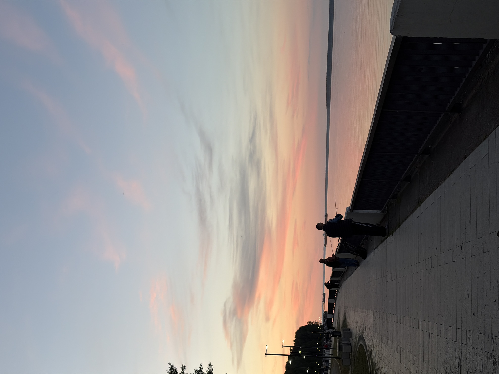 The image captures a serene sunset or sunrise over a wide body of water, likely the Kama River in Chaikovsky, Russia, as viewed from an embankment. The sky, dominating the upper half of the frame, displays a soft gradient of light blue and pale grey that transitions into warm hues of pink, orange, and yellow closer to the horizon. Scattered, wispy clouds streak across the sky, catching the vibrant colors of the setting or rising sun.
Below the horizon, the calm water reflects the subtle colors of the sky, appearing as a smooth expanse with gentle transitions of blue, pink, and purple. Along the right side of the image, a paved promenade or embankment stretches into the distance, bordered by a dark, decorative railing.
Several individuals are visible along the railing, silhouetted against the bright sky and water. They appear to be engaged in leisure activities, with at least two figures holding fishing rods, suggesting they are fishing. Their dark forms emphasize the dramatic lighting of the scene. In the mid-ground, on the far side of the water or along the distant part of the embankment, there are faint silhouettes of trees or foliage and a few streetlights, adding depth to the composition. The overall atmosphere is peaceful and contemplative. No visible text is present in the image.