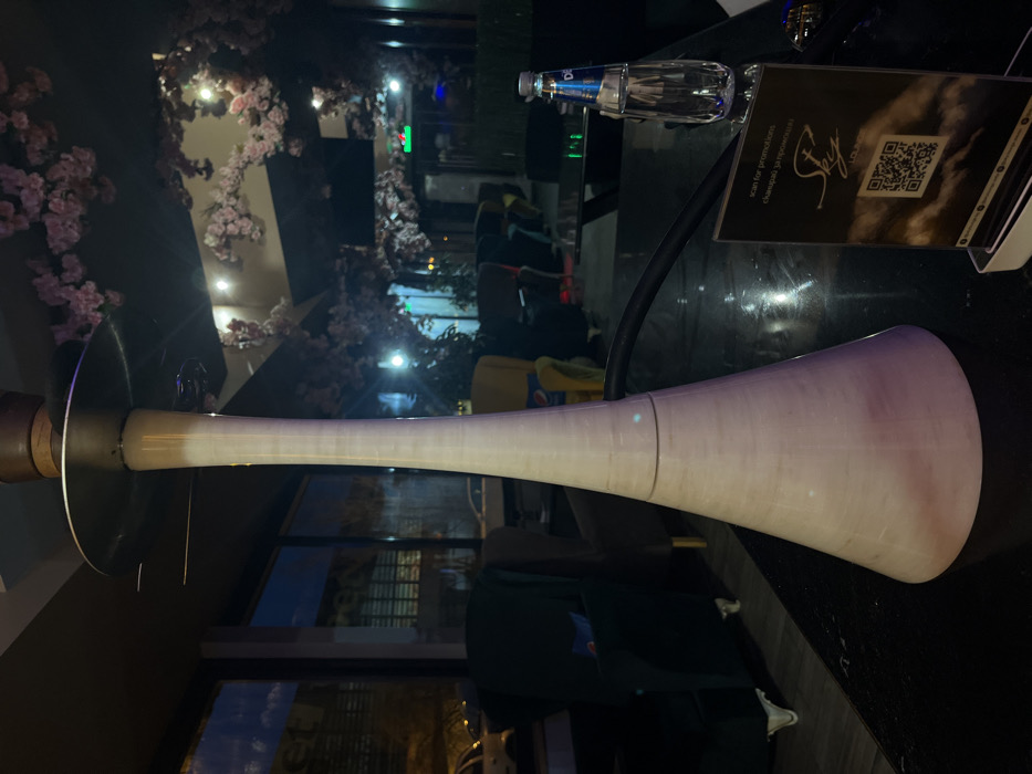 The image depicts the interior of a shisha lounge, specifically a Sky Lounge establishment located in Sofia, Bulgaria, likely at Ul. "Hristo Belchev" 3.
In the foreground, a prominent modern shisha (hookah) stands vertically on a dark, reflective tabletop. The hookah features a tall, slender, conical base and shaft, predominantly white with subtle horizontal striations, topped by a dark brown element and a metallic tray. A black shisha hose extends from its base to the right. To the right of the hookah, a transparent plastic bottle of Devin mineral water with a blue cap is positioned. Immediately in front of the water bottle, a black tabletop menu stand displays "Sky Lounge" branding, a QR code, and text in both English ("scan for promotions") and Bulgarian ("сканирай за промоции").
The background reveals a dimly lit lounge area. Large windows on the left reflect interior lighting and show blurred external elements, including the outlines of parked cars and distant streetlights, indicating an evening or nighttime setting. The interior features various seating arrangements, including dark armchairs with light-colored legs and at least one yellow armchair visible further back. The ceiling and upper walls are extensively decorated with artificial pink cherry blossoms, creating a thematic ambiance. Additional warm light sources are visible within the ceiling and reflecting off the walls. No individuals are visible, and no direct actions or interactions are depicted.