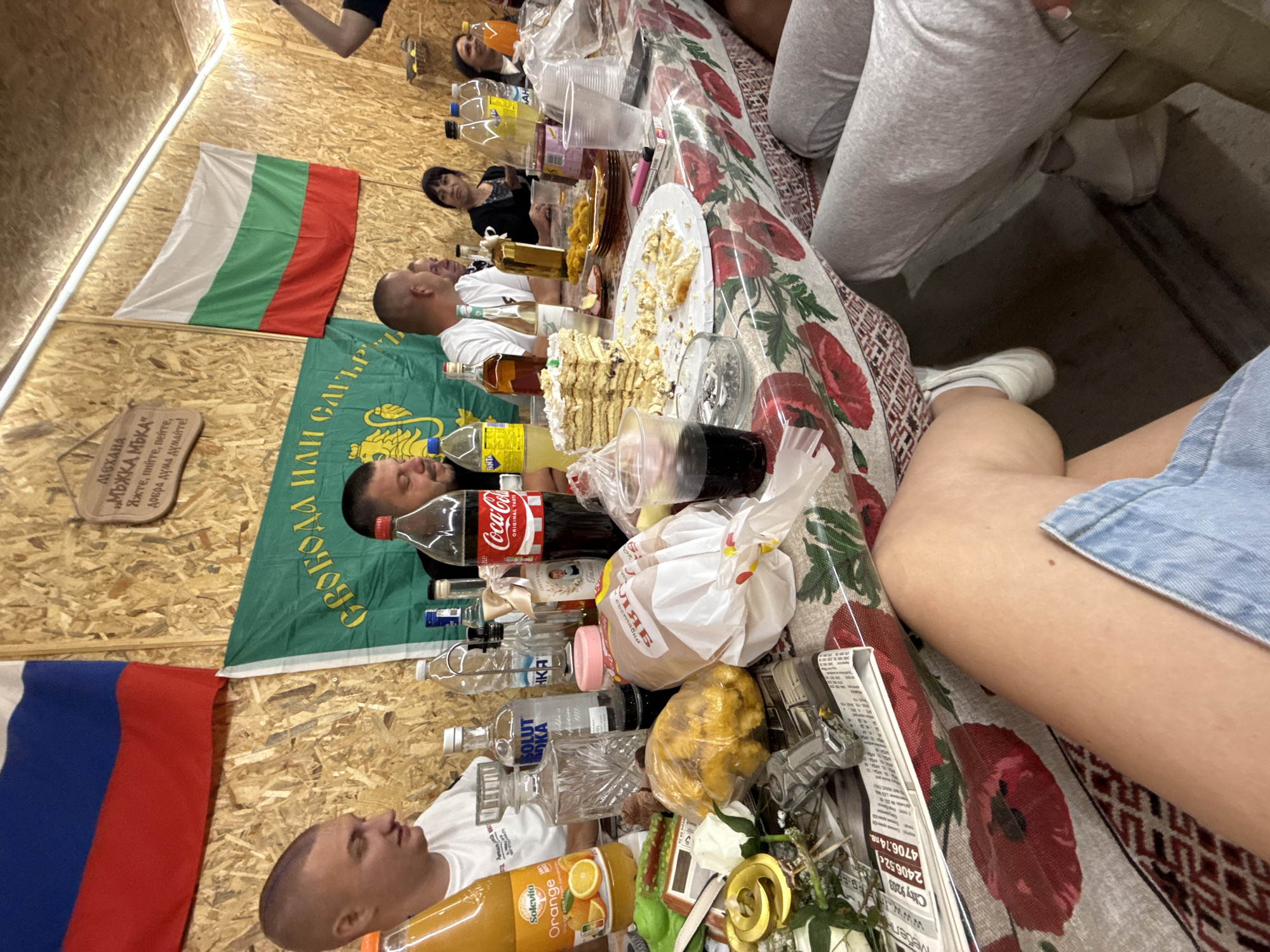 A group of people are gathered around a table laden with food and drinks, likely celebrating an event. The setting appears to be outdoors or in an open-air venue, with wooden walls and ceiling visible. Flags of Russia and Bulgaria are prominently displayed, suggesting a connection to both countries. The table is covered with a floral-patterned tablecloth and features a cake, snacks, bottles of alcohol, and soft drinks. Several individuals are seated at the table, engaged in conversation and consuming food and beverages. The atmosphere seems festive and informal. Based on the Bulgarian flag and the general ambiance, the location is likely the city of Rogosh, Bulgaria.