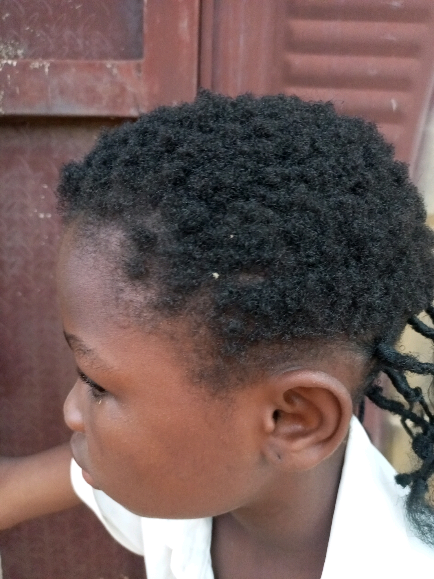 The image shows a close-up of a young child's head, from the side, in what appears to be a residential area of Tempure, Nigeria. The child has dark, curly hair styled into braids that extend down the back. The child is wearing a white collared shirt. The background consists of a reddish-brown, textured surface, possibly a wooden door or wall. The lighting suggests it is daytime.