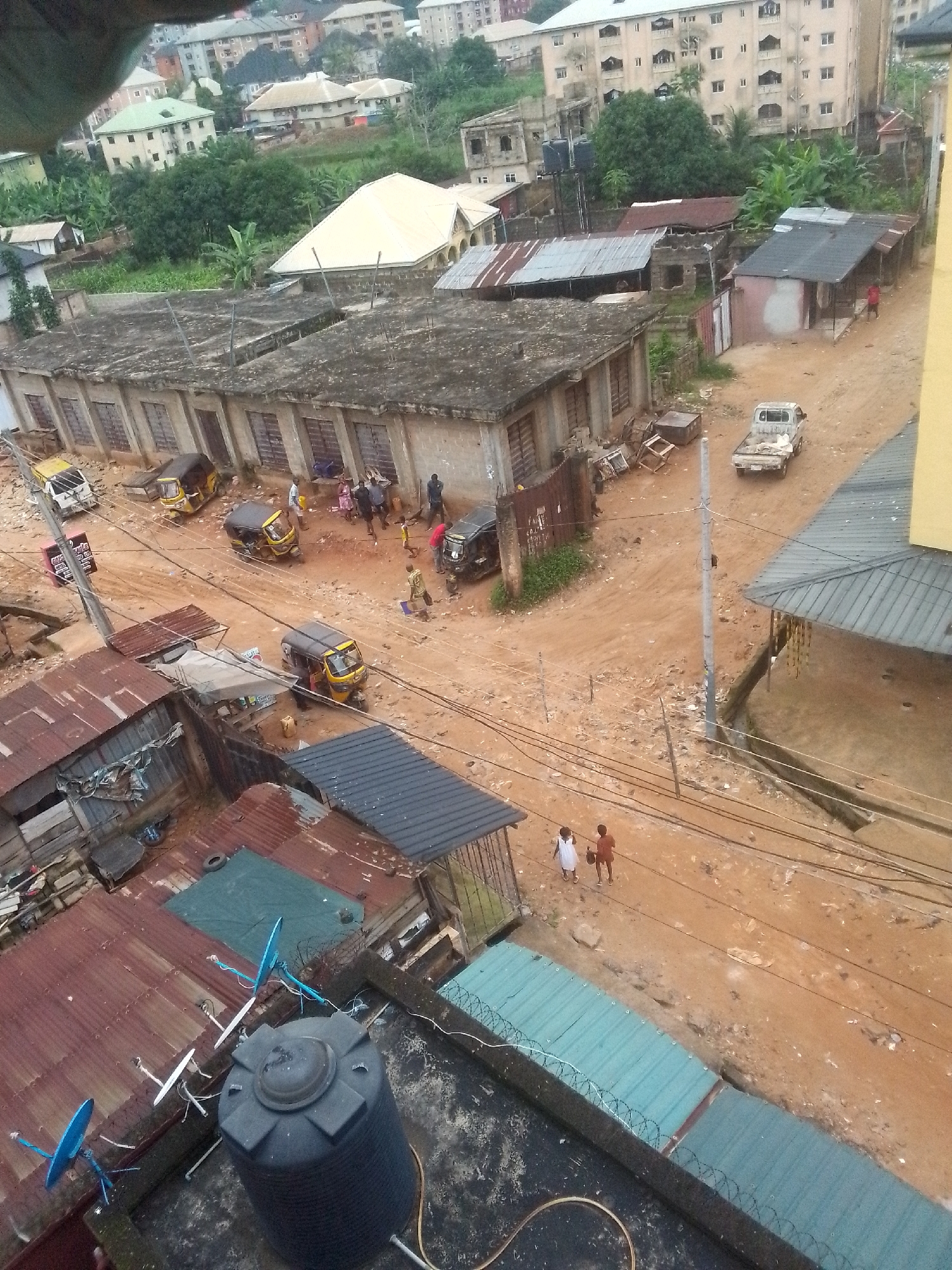 The image depicts a street scene in Nkpor, Nigeria, characterized by a dirt road and densely packed, low-rise buildings with corrugated metal roofs. In the foreground, a large black water tank sits on a rooftop, surrounded by several satellite dishes. A group of people are visible on the dirt road, some near a light-colored pickup truck, others gathered around several motorized three-wheeled vehicles, commonly known as "keke napeps" or auto-rickshaws. A few individuals appear to be interacting with the vehicles, suggesting activity related to transportation or commerce. Further back, more buildings are visible, some appearing weathered or under construction. The overall environment suggests a busy, possibly informal urban area.