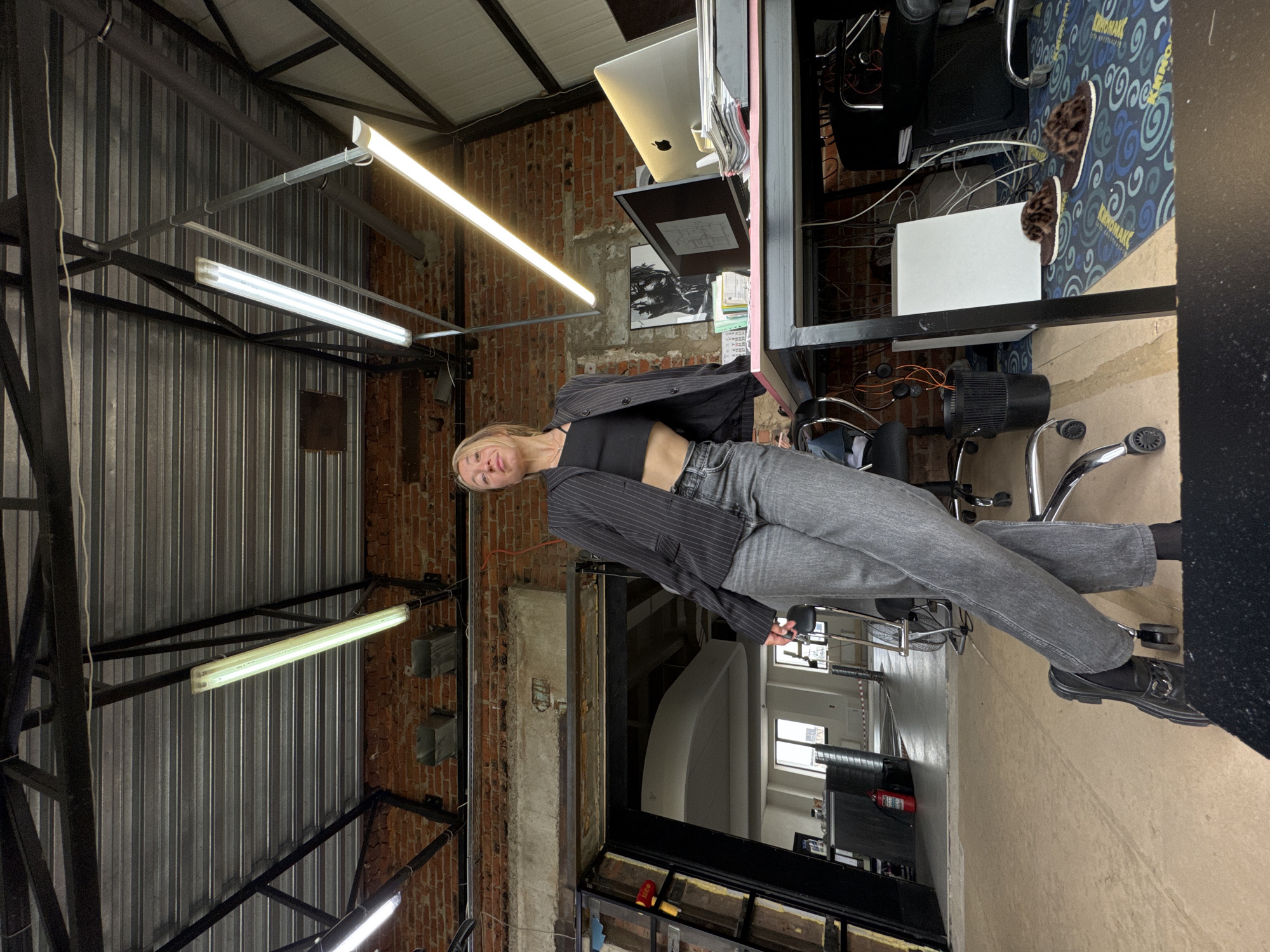 A young woman with blonde hair, wearing a black crop top, a striped grey blazer, and grey jeans, poses in an industrial-style interior. She is looking towards the camera with a neutral expression, her head tilted slightly. Her feet, clad in black socks and black loafers with a silver buckle, are visible towards the bottom right.
The setting appears to be a repurposed industrial space, characterized by exposed red brick walls and corrugated metal panels that serve as both walls and ceiling elements. The ceiling is supported by dark metal beams and features multiple bright fluorescent tube lights, casting illumination over the scene.
To the woman's right, there is an office-like setup with a silver Apple iMac and another computer monitor visible on a surface. Wires and cables are noticeable around this area, along with several office chairs with wheels. On the floor, a blue and green patterned rug with the text "KRYOMAK" is partially visible, where a pair of leopard-print slippers rests. A white box-like object is also on the floor near the rug. In the background, partially obscured, a red fire extinguisher can be seen. A framed piece of art hangs on the brick wall behind the desk area. The overall ambiance is modern and urban.