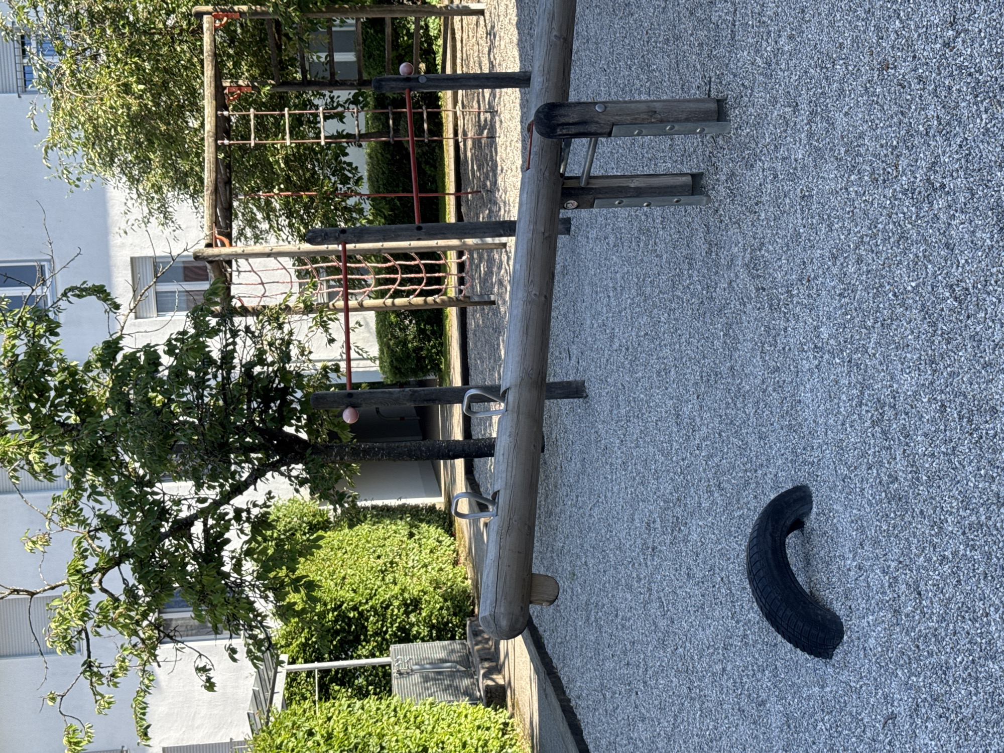 The image depicts a playground scene in Höhenkirchen-Siegertsbrunn, Germany, likely during daytime given the bright lighting and shadows. The setting is an outdoor playground with a gravel surface.
Prominent objects include a wooden seesaw, part of a climbing frame with ropes and wooden rungs, and a detached black tire lying on the gravel. The climbing frame appears to be constructed from wooden poles and red ropes, with some metal supports. The seesaw is made of a thick wooden log with metal handles for users. The surrounding area is lush with green trees and hedges, suggesting a well-maintained outdoor space. In the background, a modern white building with windows is visible, indicating a residential or institutional area adjacent to the playground.
No people are visible in the image, so no activity or event is explicitly taking place. The image is framed to focus on the playground equipment and the surrounding environment. The lighting suggests it is a sunny day, casting distinct shadows on the gravel. No text is visible in the image. The overall impression is one of a quiet, perhaps unused, playground.