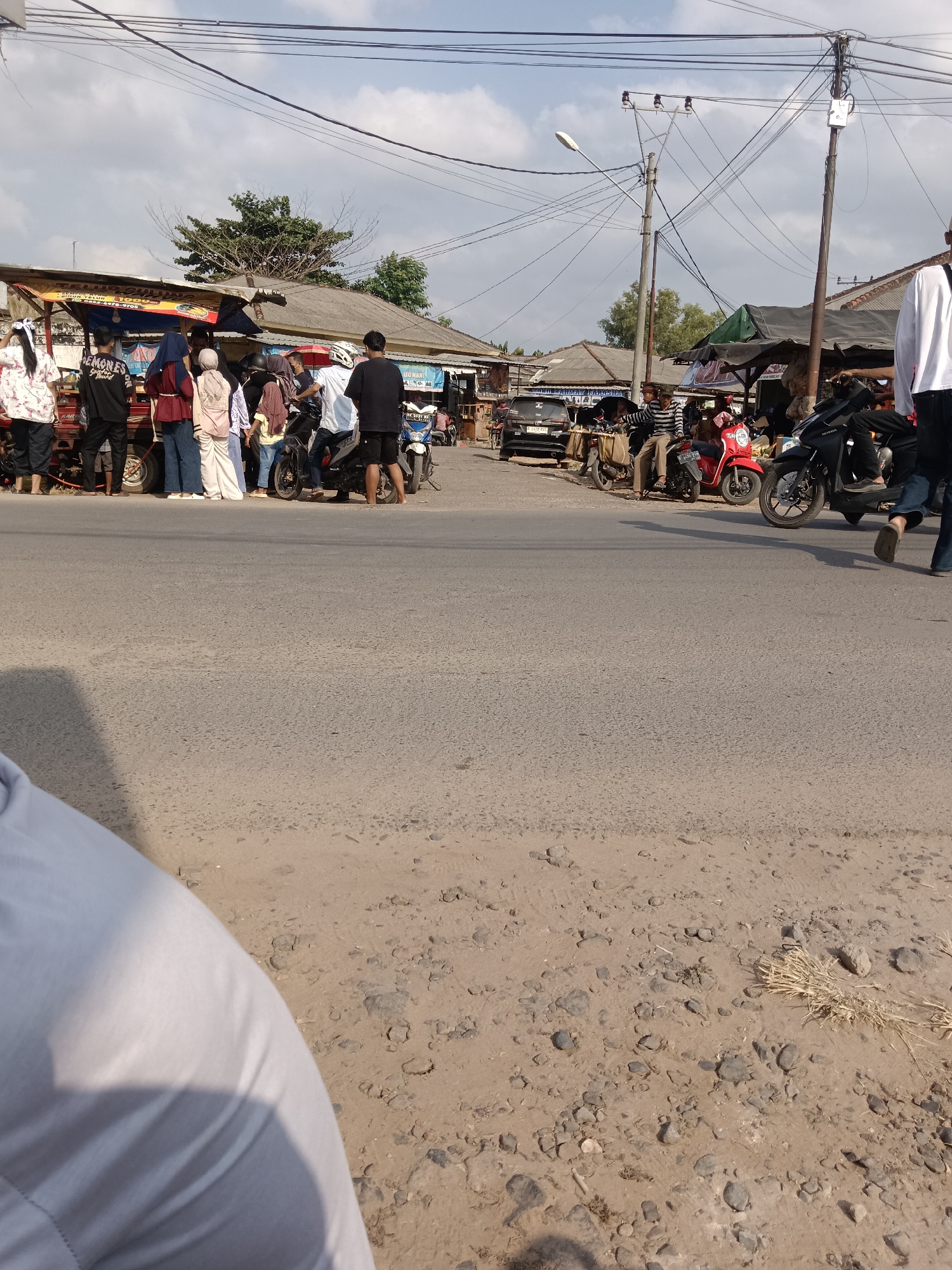 A street scene in Tegal Gondo, Indonesia, shows a group of people gathered around several food stalls and parked motorcycles. In the foreground, a person's arm, clad in a white shirt, is visible, casting a shadow on the dusty ground. Beyond this, a paved road leads to the activity. Several food stalls are set up along the side of the road, with awnings providing shade. People are standing in line, some are seated on motorcycles, and others are interacting. Motorcycles are parked in a row, suggesting a busy commercial area. The background features more stalls, buildings, utility poles with a complex network of wires, and trees under a partly cloudy sky. The overall atmosphere appears to be a bustling local market or street food hub.
