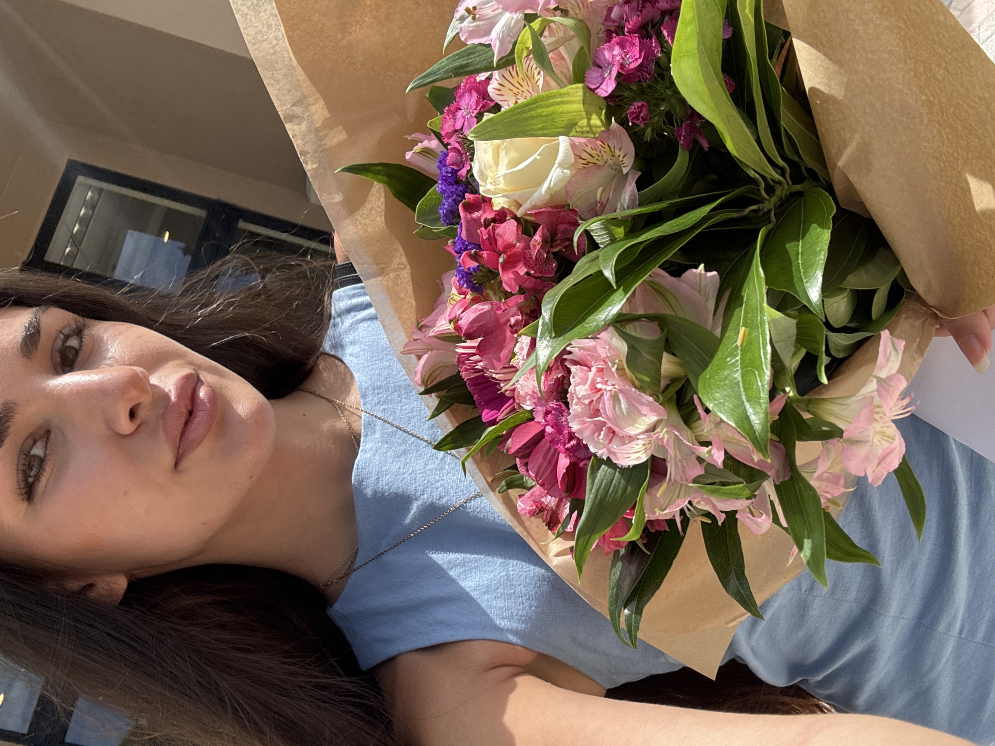 A young adult female is depicted in a sunlit outdoor setting, holding a substantial mixed floral bouquet wrapped in brown kraft paper. She has long, dark brown hair, brown eyes, and faces the camera with a subtle smile. She is wearing a light blue sleeveless top and a thin gold-colored chain necklace.
The bouquet consists of various flowers, prominently featuring a white rose, pink and fuchsia alstroemeria, deep pink dianthus, and small purple blossoms (likely limonium), surrounded by green foliage. The brown paper wrapping exhibits folds and creases. Her right hand is partially visible at the bottom right, supporting the bouquet.
The background is out-of-focus, showing a light-toned building exterior with a dark-framed window or door. Strong shadows are cast on her clothing and the flower wrapping, indicating direct sunlight.
No specific visual cues or identifiable landmarks are present within the image to confirm the location as Boca Raton, United States of America.