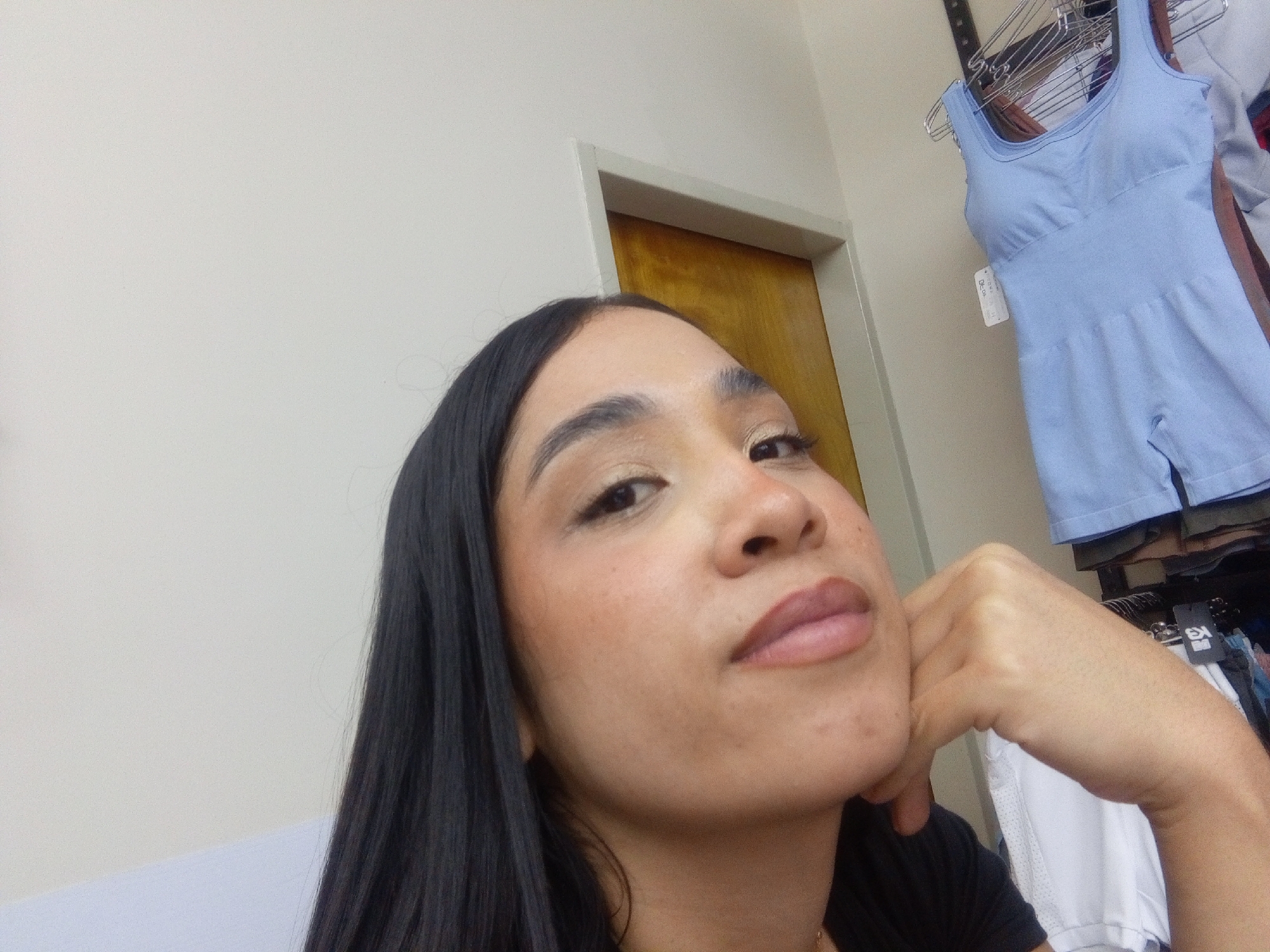A close-up image shows a woman posing for a selfie indoors. She has long, dark, straight hair and is looking slightly upwards and to the right with a neutral or pensive expression. Her left hand is resting against her chin. She appears to be wearing makeup, including eyeshadow and lip liner. Her skin has a warm undertone with some visible texture or freckles.
The background features a light-colored wall. Behind her head, a light-colored door frame outlines a wooden door. To the right of the frame, there's a clothing rack holding various garments. A light blue, seamless one-piece garment, possibly a romper or shapewear, is prominently displayed on a hanger. A small white tag with indistinct text, possibly "DE-05", is attached to the blue garment. Other clothes are visible stacked or hung beneath it. The indoor setting, with the clothing display, suggests a retail environment or a personal space with organized clothing in Valle de La Pascua, Venezuela. The lighting appears soft and ambient.