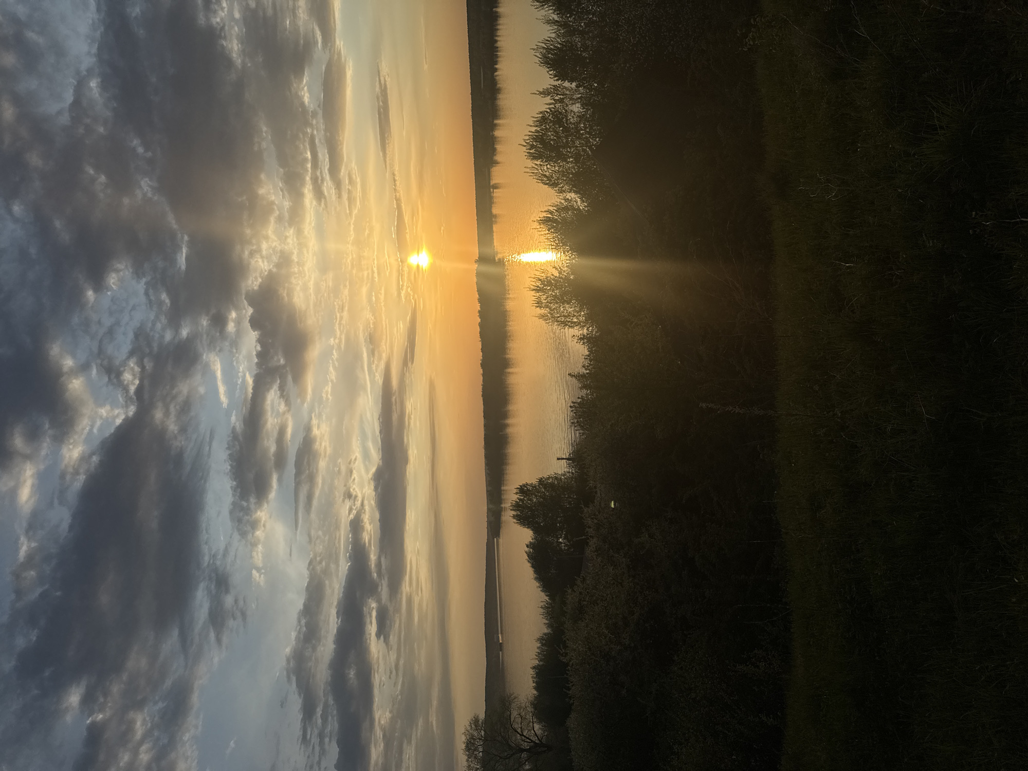 The image shows a landscape with a sunset over a body of water, likely a lake or river. The location appears to be in a natural setting with trees and vegetation along the shoreline. The sun is low in the sky, casting a golden light and creating reflections on the water's surface. Clouds are visible in the sky, contributing to the overall atmospheric effect. The location is likely near Ольховка, Russia, given the context of the image. No people or other specific subjects are visible.