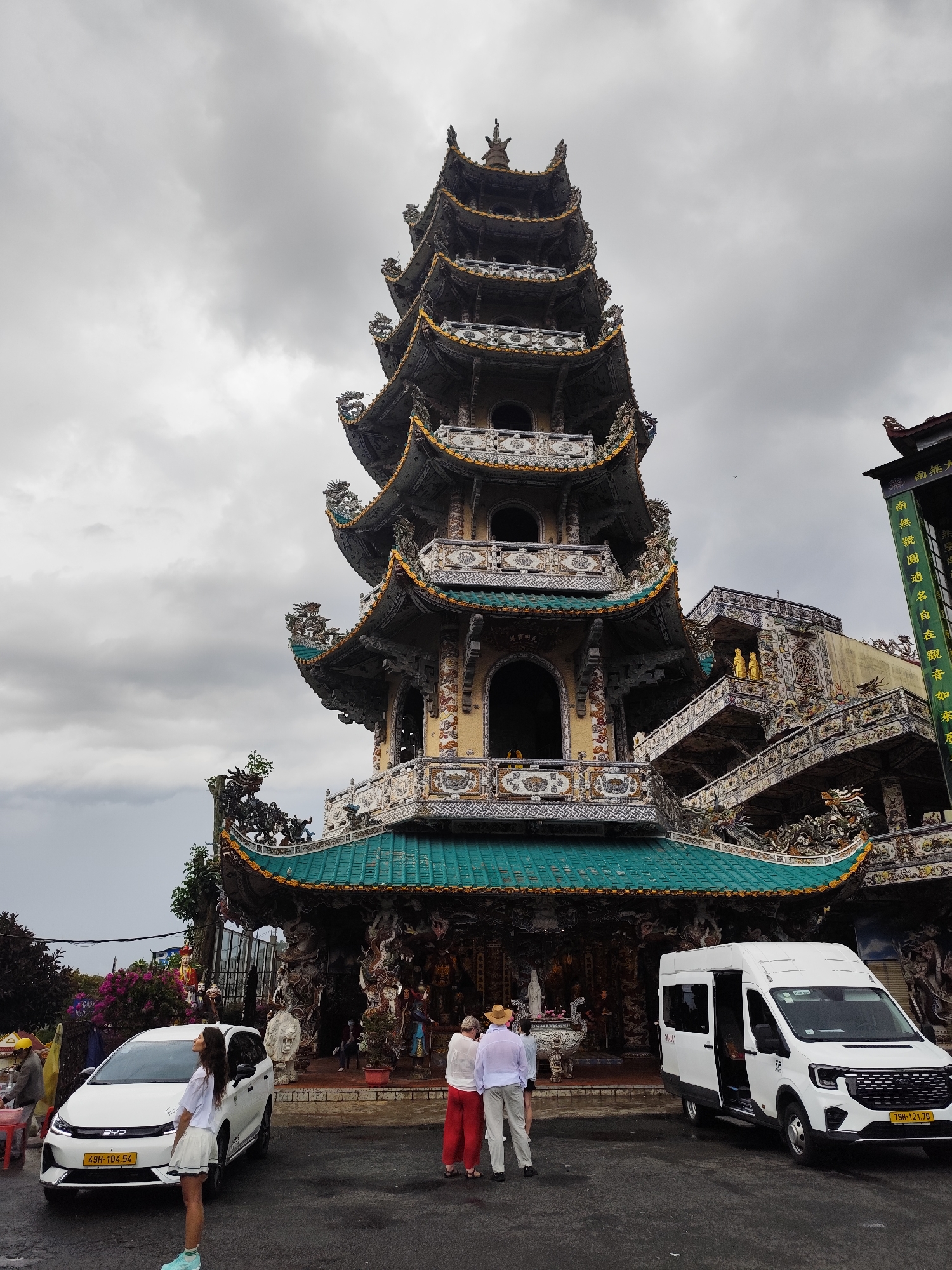 The image captures the elaborate architecture of a Buddhist temple in Xuân Thành, Vietnam. The multi-tiered pagoda is the focal point, displaying intricate designs and decorations. People are seen interacting near the base of the temple, with two white vehicles parked on either side. The sky is overcast, contributing to the overall atmosphere of the scene. The composition includes foreground elements like people and vehicles, leading the eye toward the detailed structure of the temple. The architecture showcases a blend of cultural and artistic elements, typical of Buddhist temples in Vietnam.