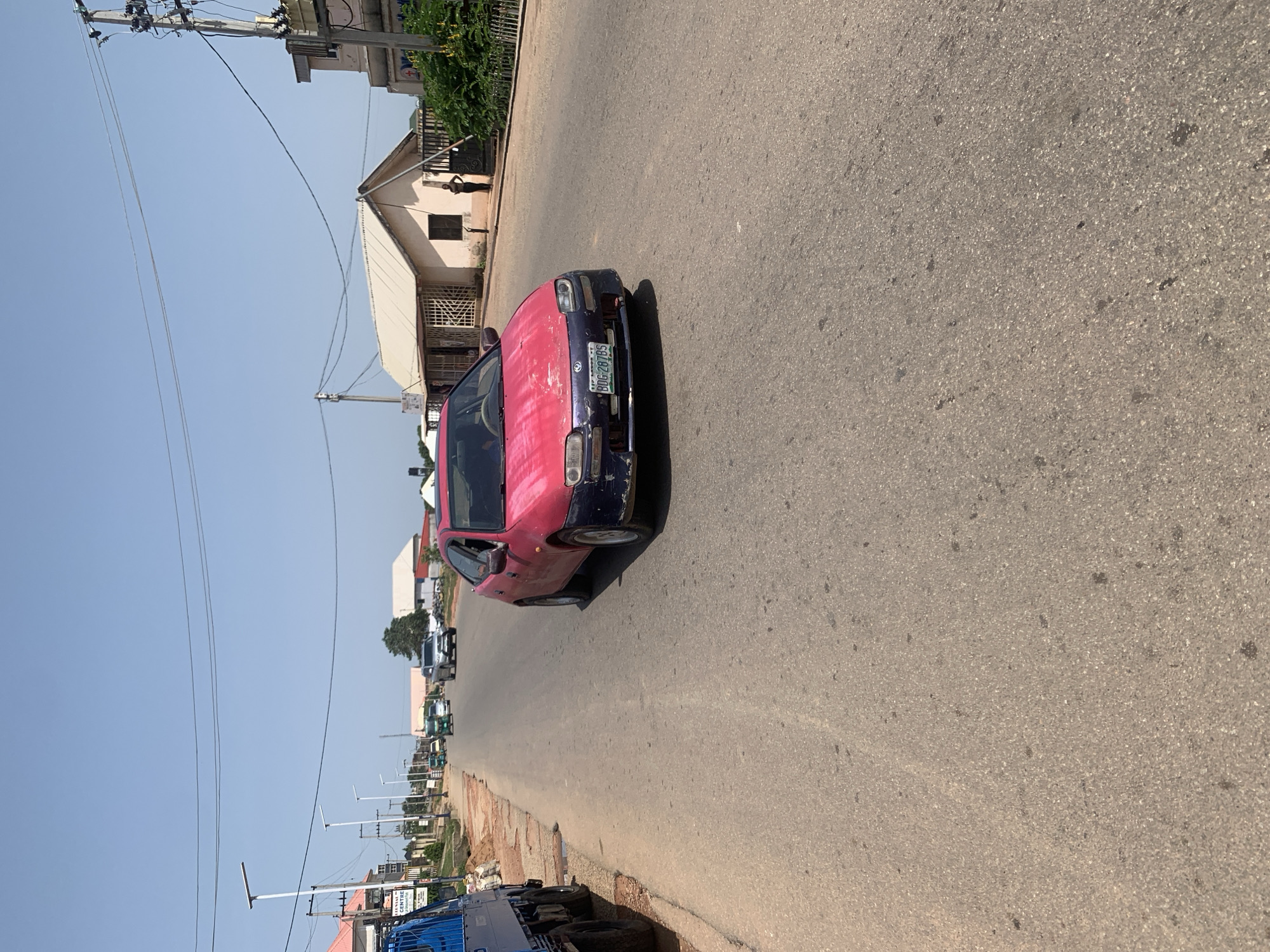 In Jos, Nigeria, on a bright, clear day, a red and blue car is seen driving down a paved road. The car, a compact hatchback, has a faded pink hood with visible wear and tear, and a blue bumper that appears damaged. Its license plate reads "BDG 287BS." Other vehicles, including a blue truck and several tricycles (also known as "Keke Napeps"), are visible further down the road. The street is lined with buildings, some with corrugated metal roofs, and utility poles with electrical wires stretch across the sky. The environment suggests a commercial or residential area within the city, with sidewalks and a mixture of construction and established structures.