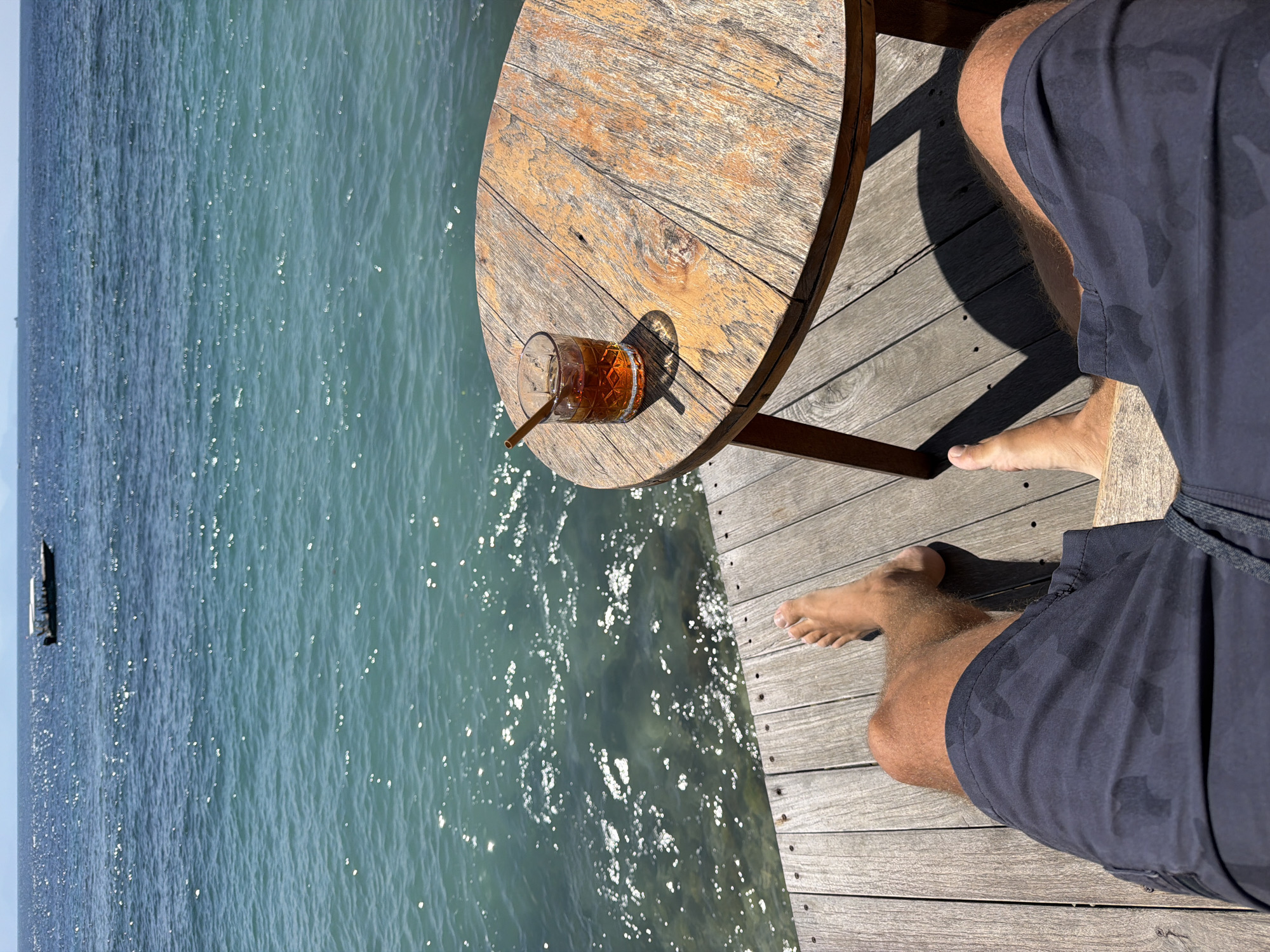A first-person perspective shot shows a person sitting on a wooden deck by the ocean. In the foreground, the person's legs and feet are visible, wearing dark, patterned shorts. A rustic wooden table sits to the right, holding a glass of amber-colored liquid with a cinnamon stick. The ocean, a vibrant turquoise, stretches into the distance, with a small boat visible on the horizon. The sun glints off the water's surface, creating a sparkling effect. The scene suggests a relaxed atmosphere in a tropical setting, likely in Nusa Penida, Indonesia, known for its beautiful beaches and ocean views.
