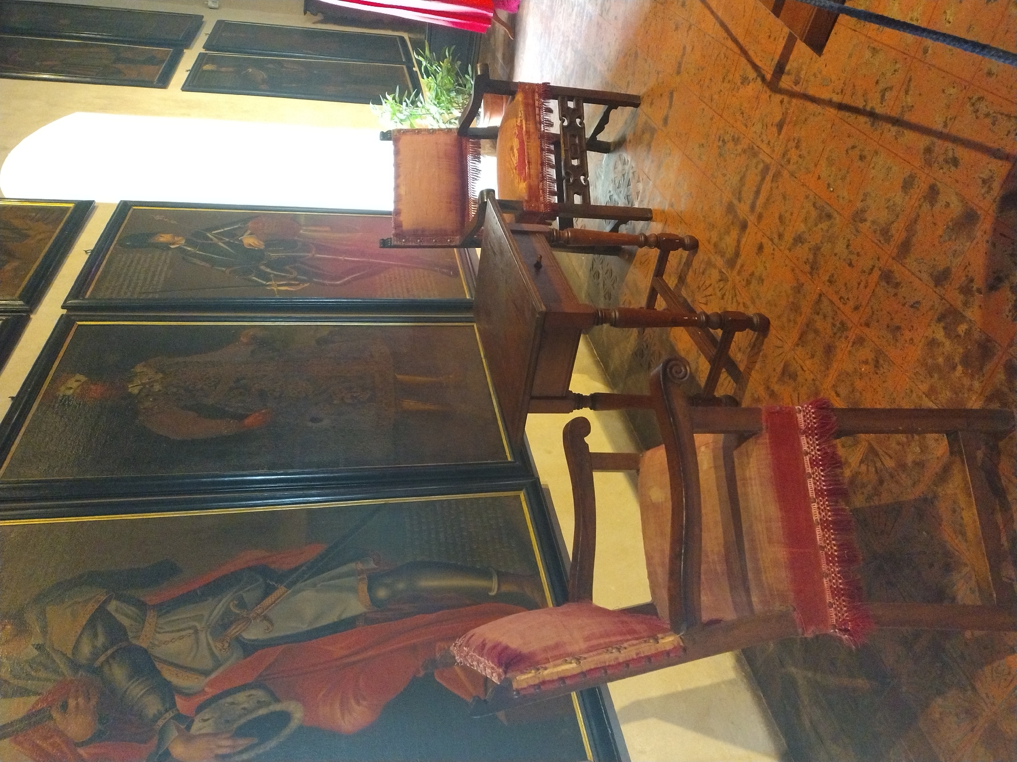 This image captures a room in the Karlštejn Castle in Karlštejn, Czechia, featuring a collection of historical paintings and antique furniture. The paintings, displayed on the wall, are large-scale portraits of individuals in period attire, one notable for its depiction of a figure in red and gold with a sword. Two wooden chairs with red upholstered seats and backs are positioned in the foreground and mid-ground. The chair on the left is closer to the viewer, while the chair on the right is further back, adjacent to a dark wooden table with a drawer. The floor is tiled with a pattern of terracotta-colored squares. Sunlight streams in from an arched window on the right side of the image, illuminating the scene and casting shadows. A potted plant is visible to the right of the window, and a hint of red fabric, possibly from a curtain or garment, is present on the far right edge.