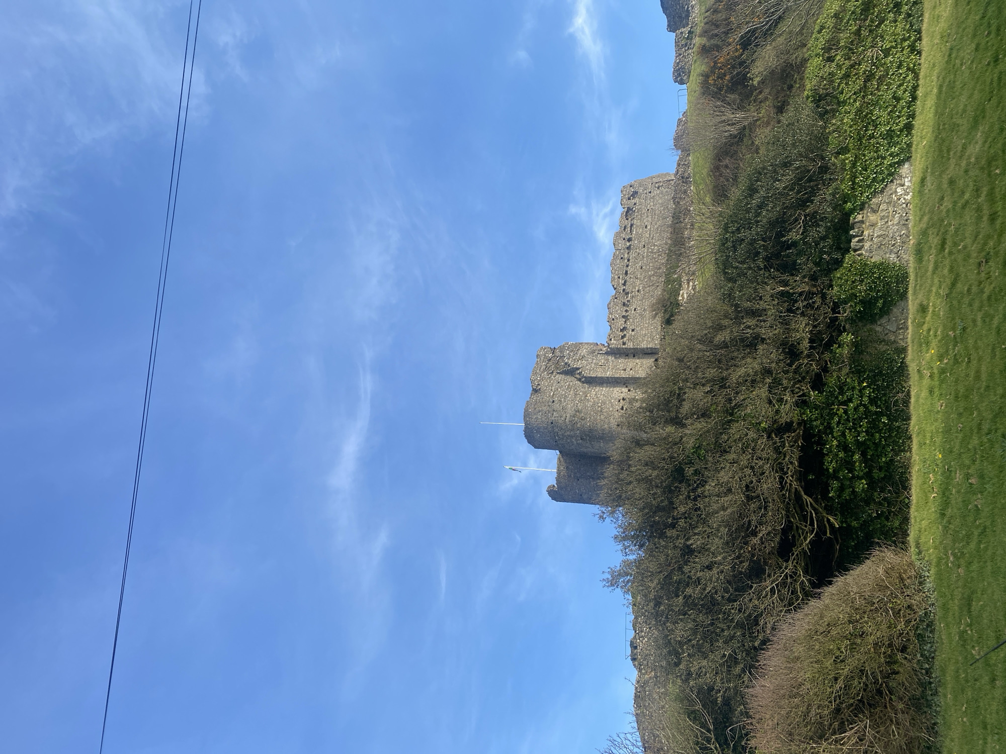 The image features Criccieth Castle, a medieval fortress in Criccieth, Wales. The castle, constructed of stone, sits atop a grassy hill and is partially obscured by trees and foliage. Two towers are visible, each topped with a flagpole. The sky is blue with thin, wispy clouds. To the left are overhead wires.