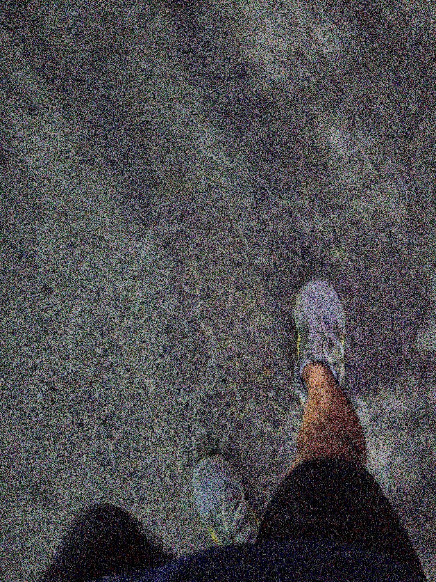 The image shows a first-person perspective of someone's legs and feet as they walk on a paved surface. The person is wearing athletic shoes, one of which is a light grey with yellow accents. The ground appears to be asphalt or concrete, with some slight texture. The lighting suggests it might be dusk or an overcast day. There are no other people or objects visible in the frame. The image quality is poor due to significant digital noise, obscuring finer details. The location in the city of Bekasi, Indonesia, is not discernible from the visual information present in the image.