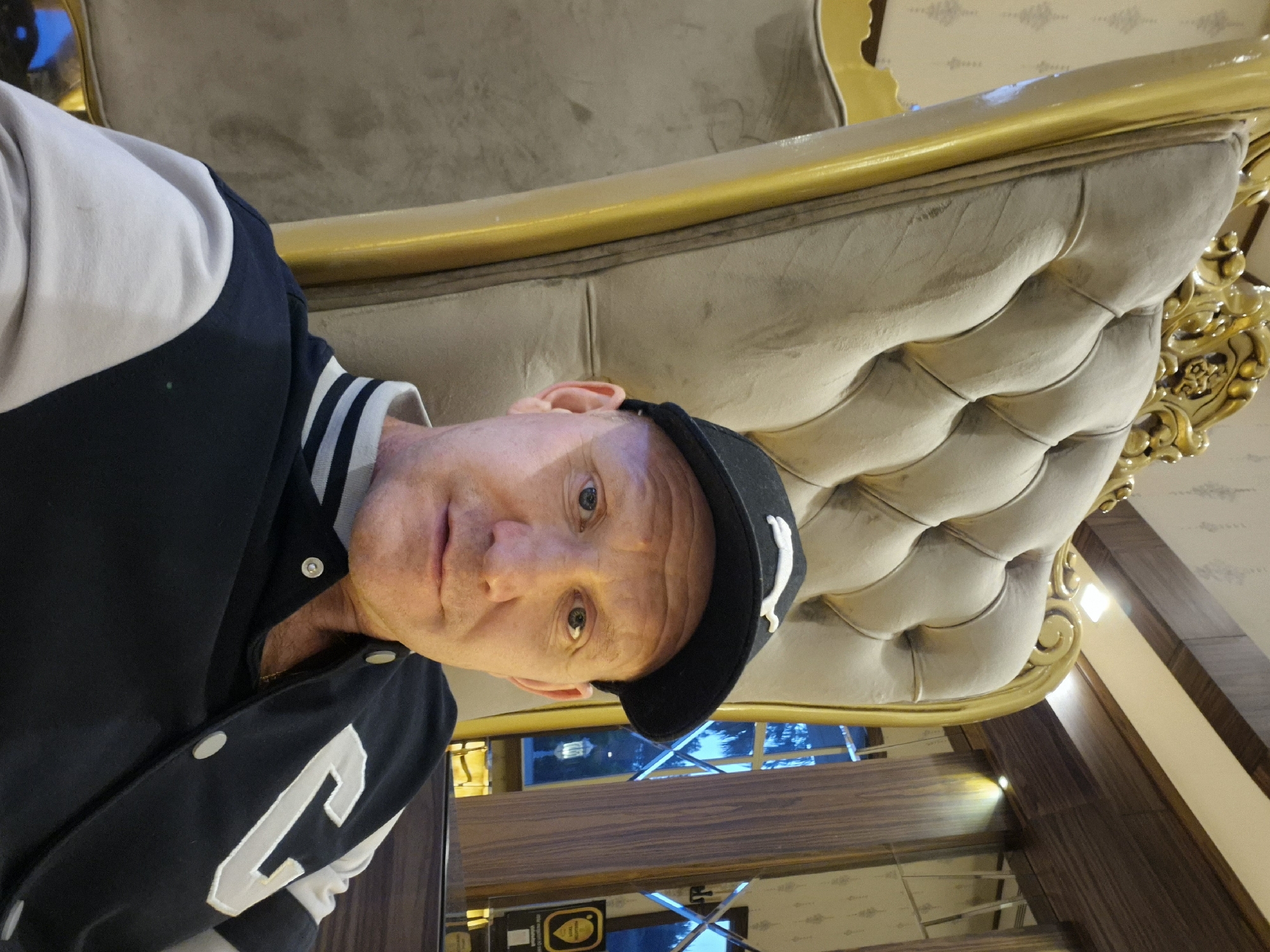 A man sits on an ornately designed, beige-colored sofa with gold trim. The sofa's structure and design are prominently displayed. The man wears a black baseball cap with a white logo, a black bomber jacket with white accents, and a striped shirt. The background features a window offering a glimpse of Alanya, Turkey, indicated by the presence of identifiable landmarks. The location is confirmed based on the distinctive architectural styles, interior design elements, and the observed outdoor scenery. A wooden wall panel and flooring are also visible.