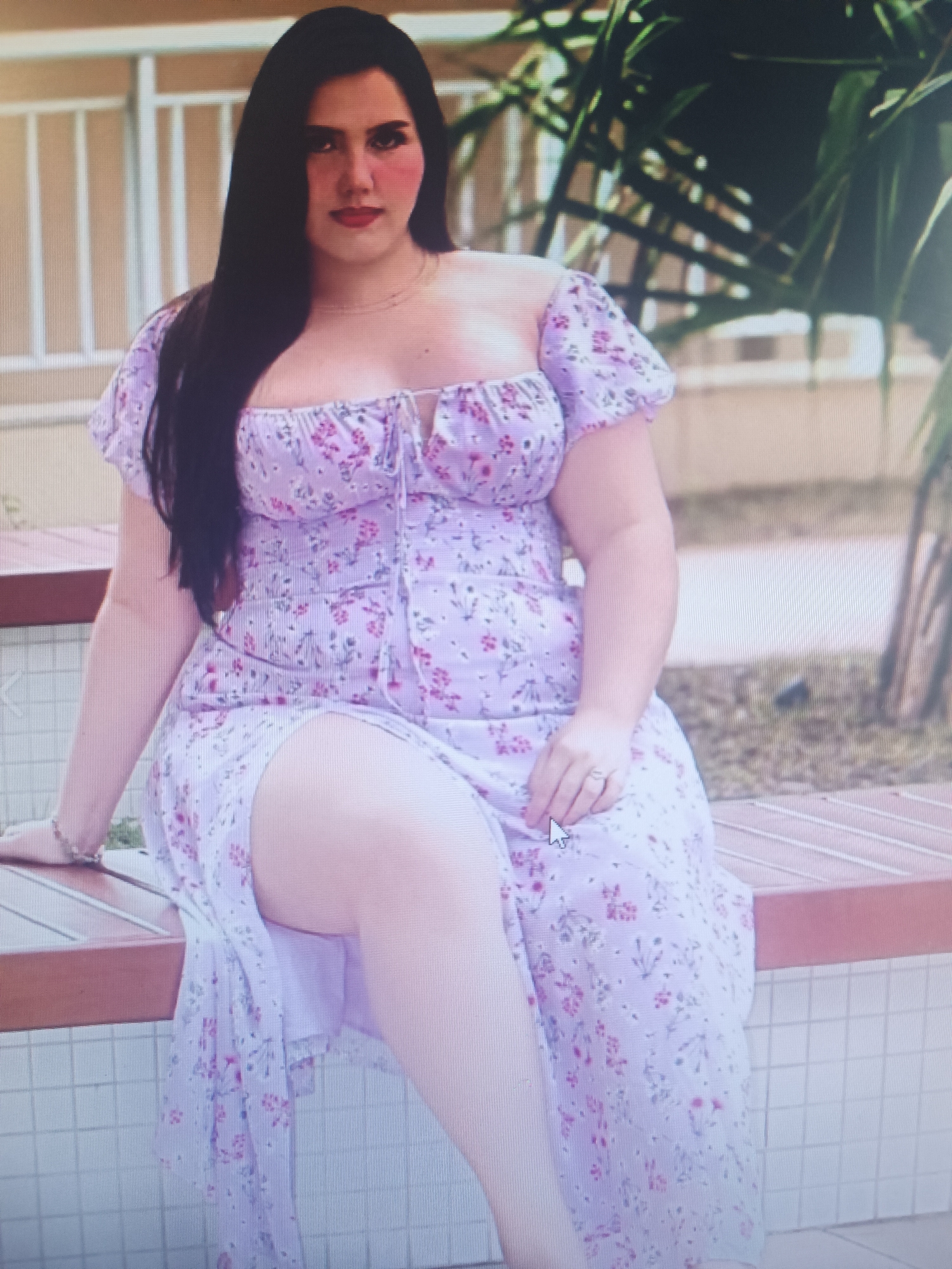 A medium shot captures a woman from the chest down, seated on a wooden bench with a tiled wall behind her. She is wearing a light purple, off-the-shoulder dress with a floral pattern of pink and green flowers. The dress has short puff sleeves and a tie-front detail at the bust. A slit in the dress reveals her leg. Her left arm rests on the bench, adorned with a bracelet. Her right hand is visible, with a ring on her ring finger. The background shows a white railing and greenery from a palm tree. The woman has long, dark hair and is wearing makeup. The scene appears to be outdoors, possibly in a park or courtyard. There is no discernible information in the image that links it to the city of Klyuchi, Russia.