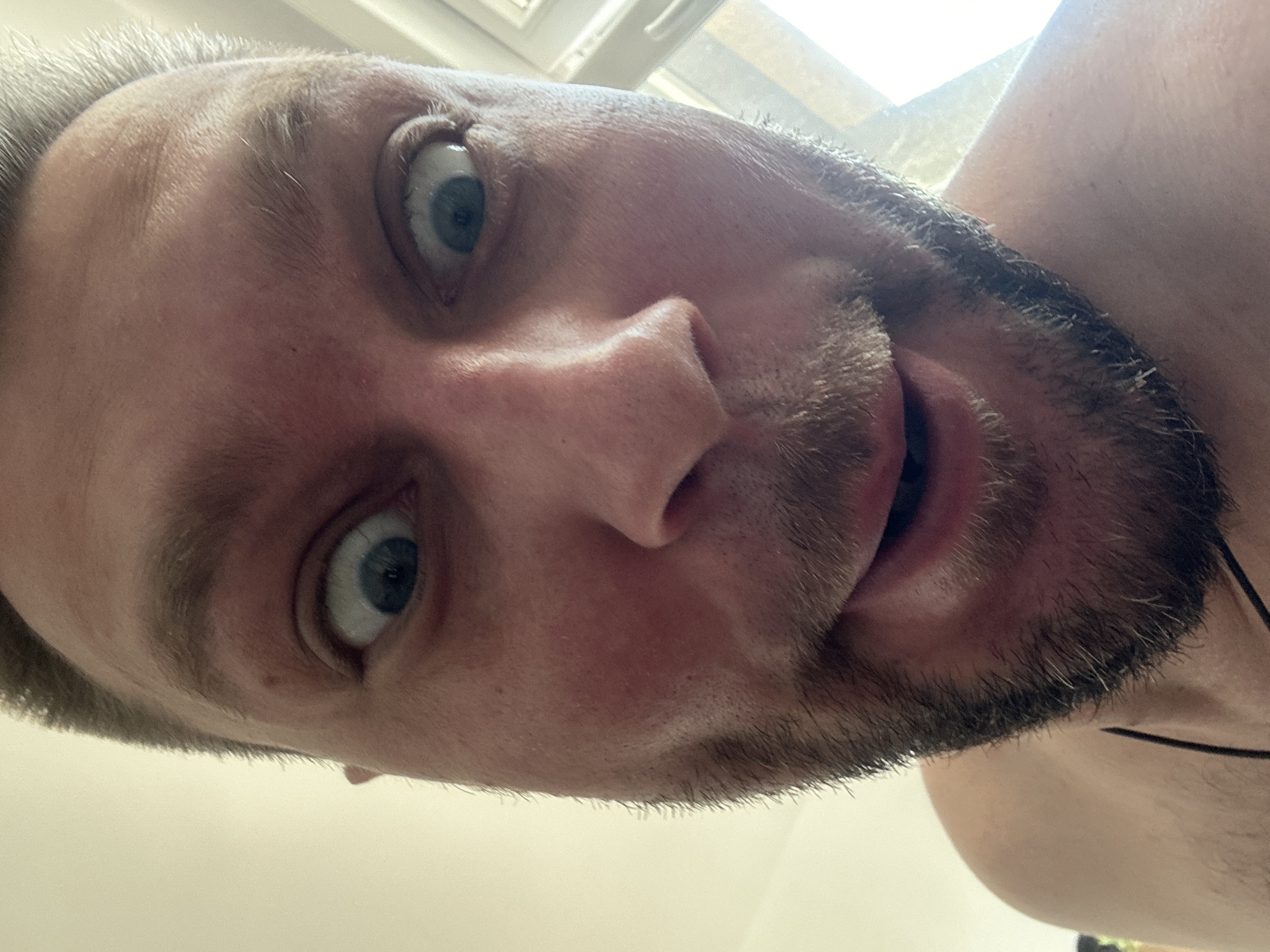 A close-up, low-angle, selfie-style photograph shows a man's face from the chest up. The man has light skin, light brown hair, and blue eyes that are wide open, giving him a surprised expression. He has a light beard and mustache. His mouth is slightly open. The background is blurred and appears to be an indoor environment with natural light coming from the right side of the frame. The image does not contain any identifiable landmarks or elements that would allow for specific location determination within Zvezdara Urban Municipality, Serbia.