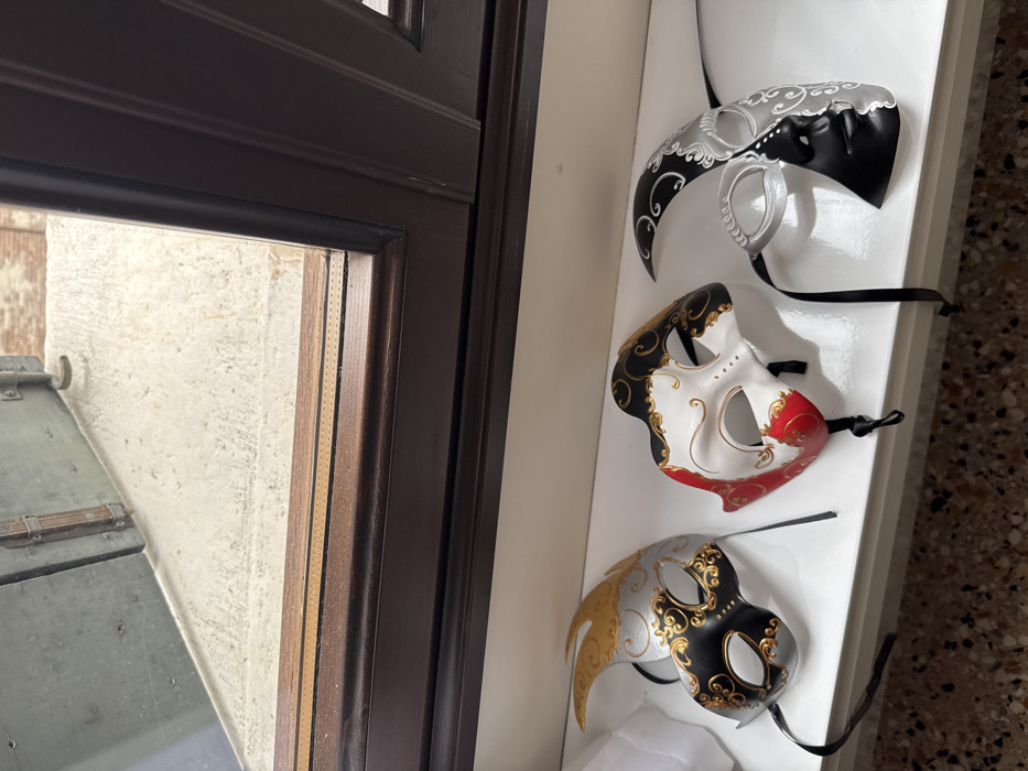 The image captures an interior view looking out a window in Venice, Italy. In the immediate foreground, three decorative Venetian-style carnival masks are displayed on a white, flat ledge. From left to right: the first mask is silver and black with gold scrollwork, featuring an elongated, curved point extending upward on the left side; the second mask is predominantly white, black, and red, also adorned with gold scrollwork; the third mask is silver and black, sculpted as a half-face with white scrollwork, depicting facial features on the black half and a crescent shape above the silver eye. All masks have black ribbons attached, which are arranged beneath them.
Below the white ledge, a section of a light brown and beige terrazzo-style floor is visible. Above the masks, a dark brown wooden window frame occupies the mid-ground, featuring a narrow, lighter brown strip with a dotted pattern along the bottom edge of the glass pane. Through the window, an exterior scene is visible. A textured, light-colored concrete or rough stone surface, likely part of an exterior ledge or balcony, is prominent. To the left, a portion of a dark green or gray wooden shutter with visible metal latch hardware is present. In the upper background, beyond the exterior surface, faint reddish-brown brickwork is discernible, consistent with Venetian architectural styles.