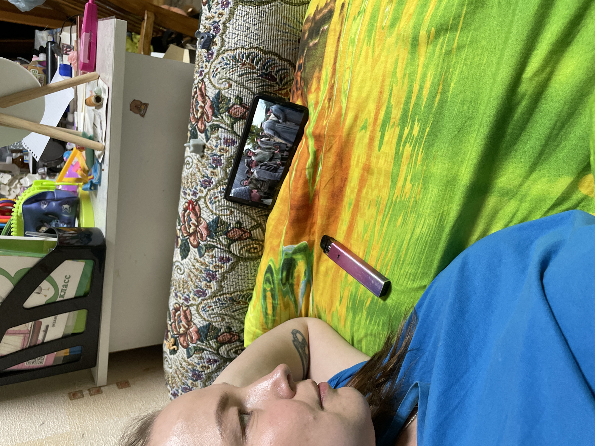 The image captures a close-up, high-angle view of a person lying down indoors, likely on a sofa or bed, in Izhevsk, Russia. The person is wearing a blue top, with their head resting on a patterned fabric that resembles a couch or an ornate blanket. Their eyes appear to be closed or looking slightly upwards, and their expression is neutral, suggesting relaxation or contemplation. A tattoo is visible on their left upper arm.
Next to the person, on a vibrantly colored green and yellow patterned blanket or pillow, rests a purple and pink metallic vape pen. A black smartphone is placed face-up on the patterned fabric, displaying what appears to be a scene from a movie or TV show. The screen shows a group of men, possibly soldiers or laborers, dressed in older-style clothing.
To the left of the person, a cluttered area featuring a white shelf or cabinet is visible. Various items like papers, a pink hair clip, and other household objects are strewn about. A black magazine or document holder on the shelf contains items with visible Cyrillic text, including the word "Класс" (Klass - Class) and fragments of "ЭКЗАМ" (Ekzam - Exam), confirming the Russian context provided. The floor beneath the shelf has a light-colored tiled pattern. The overall setting is domestic and appears somewhat lived-in, under ambient indoor lighting.